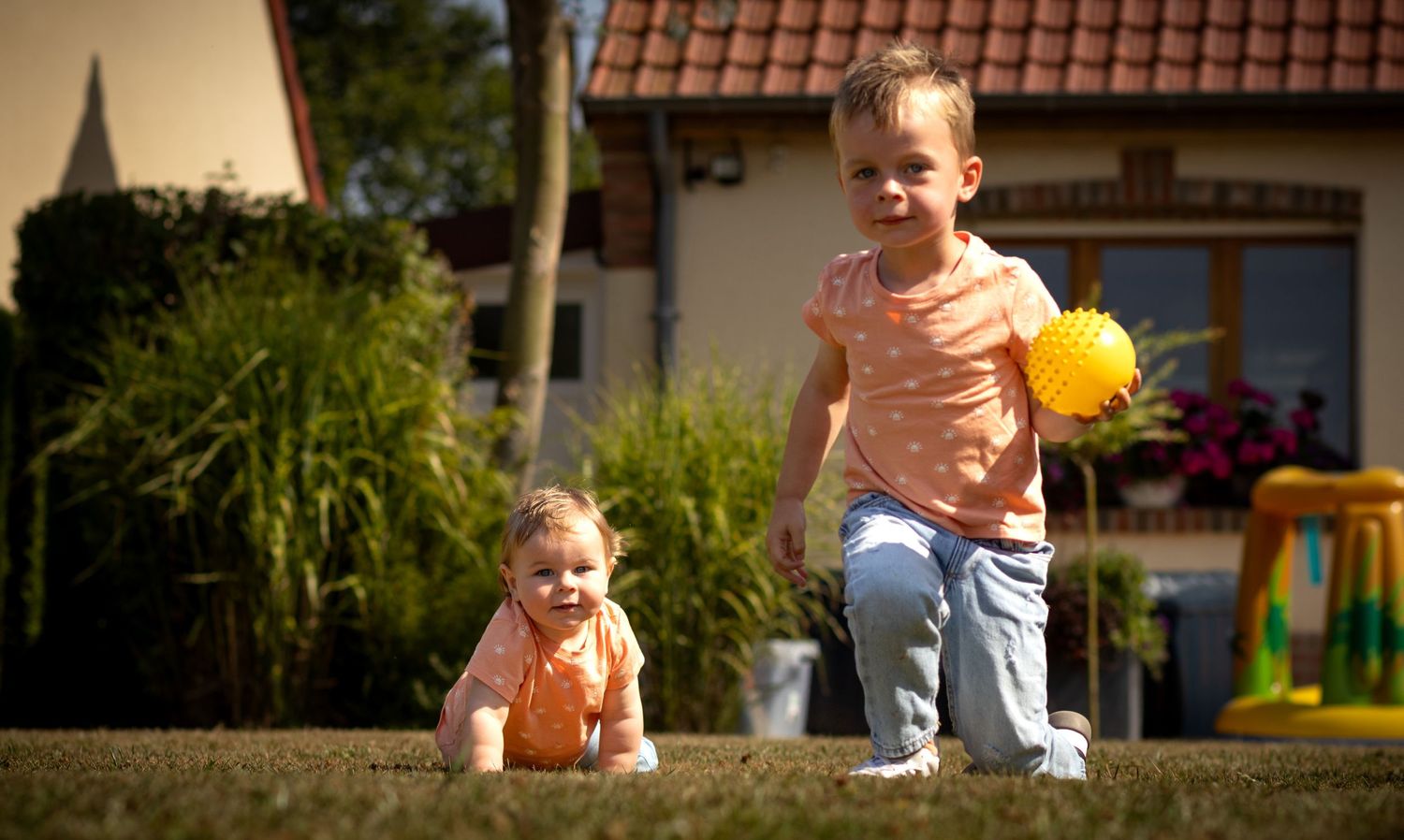 Two young children playing outdoors on grass, one crawling and one walking with a yellow ball on a sunny day.