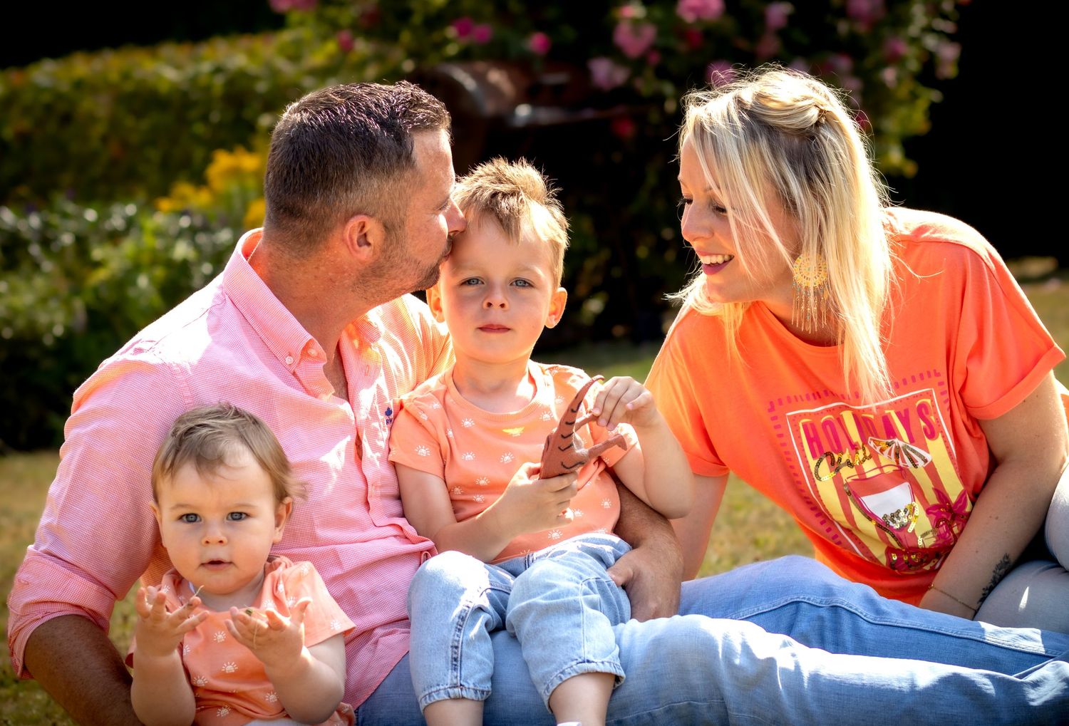 Happy family of four sitting outdoors in sunny garden, parents in pink and orange shirts with two young children.