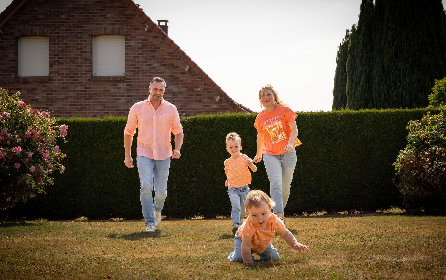 Happy family in matching orange outfits playing on green lawn beside brick house and tall hedges on sunny day.