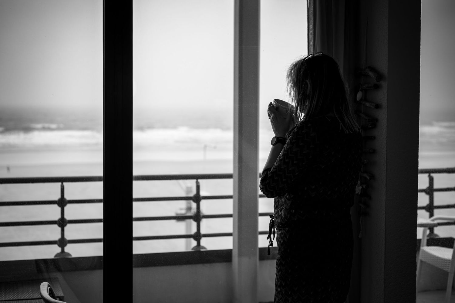 Black and white photo of a woman holding a cup, gazing out a balcony window overlooking the ocean.