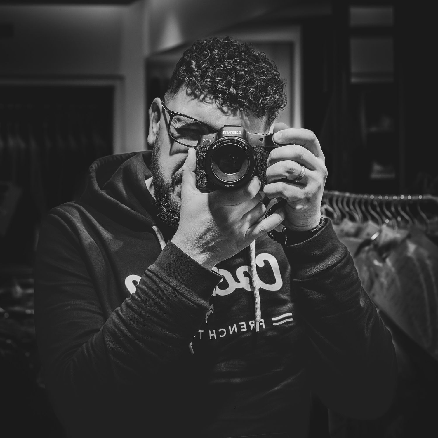 Black and white photo of a man with curly hair and glasses holding a camera up to his face in a dark room.