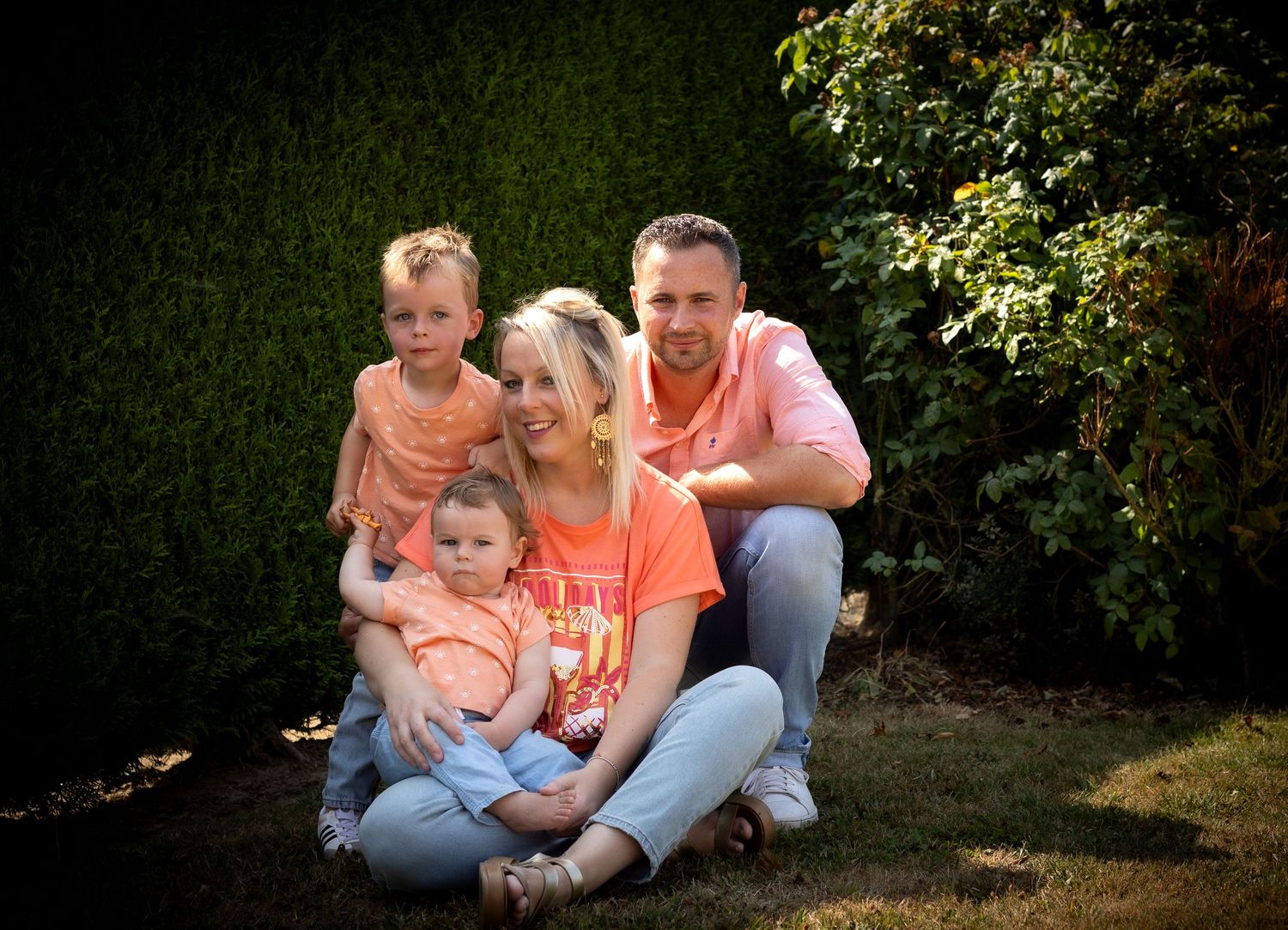 Family of four posing outdoors in matching coral outfits, with two young children, sitting on green grass near hedges.