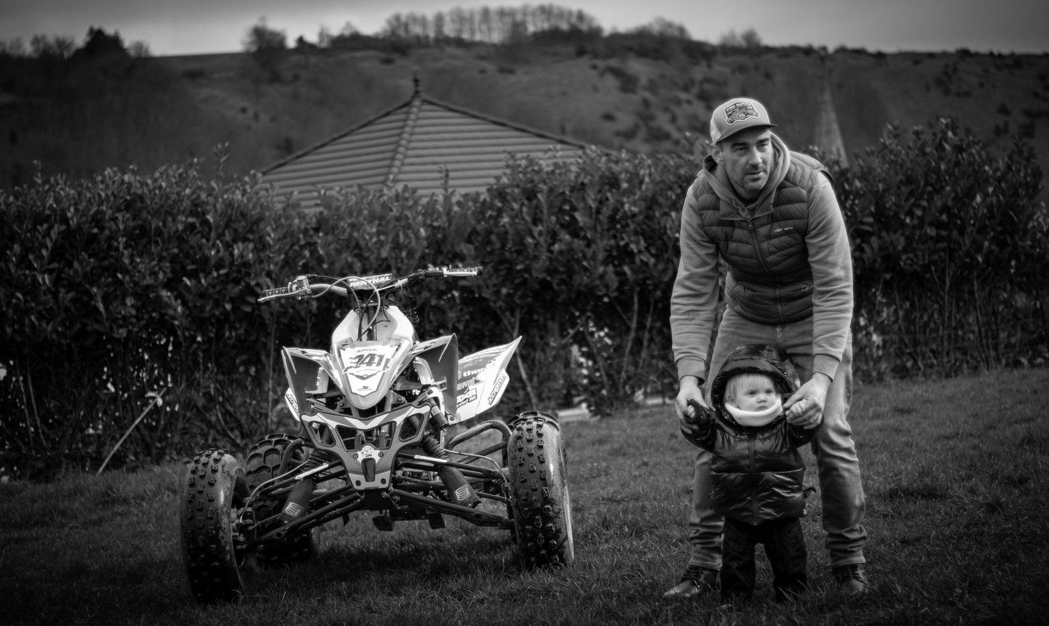 Black and white photo of an ATV parked in a rural field with someone standing beside it in winter gear.