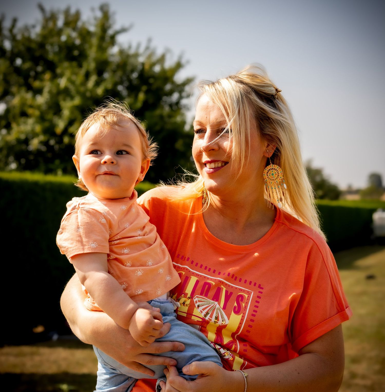 Smiling blonde woman in orange shirt holding a happy toddler outdoors on a sunny day.