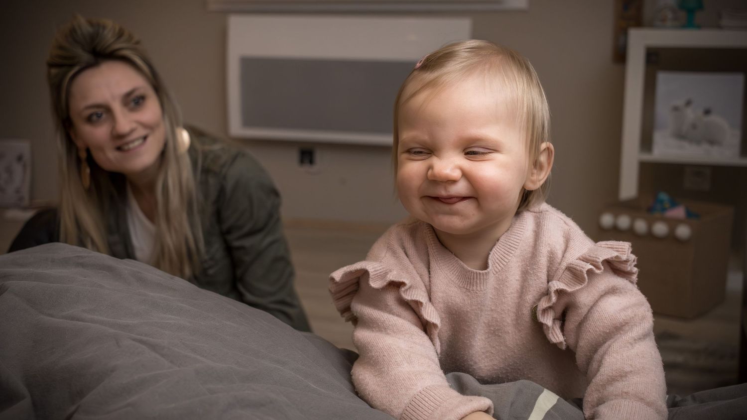 Smiling baby girl in pink ruffled outfit on bed with blonde mother watching fondly in soft-lit nursery.