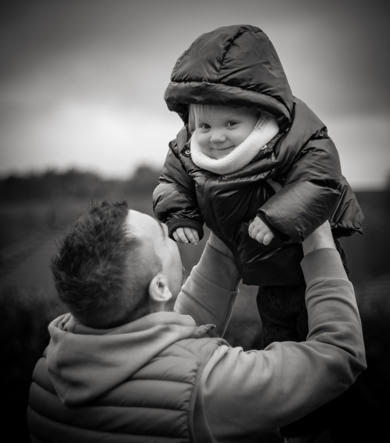 Black and white photo of a father lifting his smiling baby in a winter jacket outdoors.