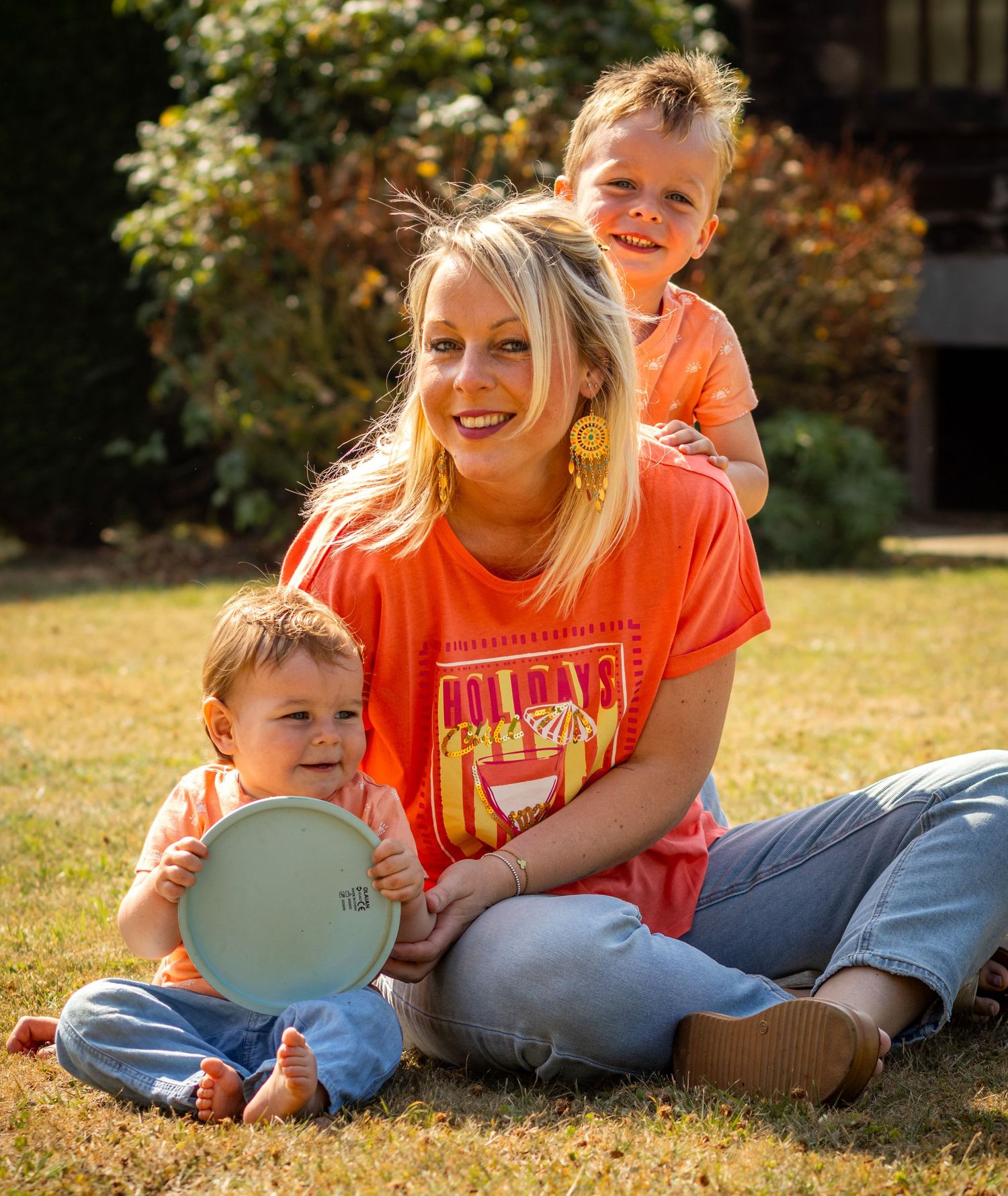 Woman in orange shirt sits on grass smiling with two young children on a sunny day outdoors.