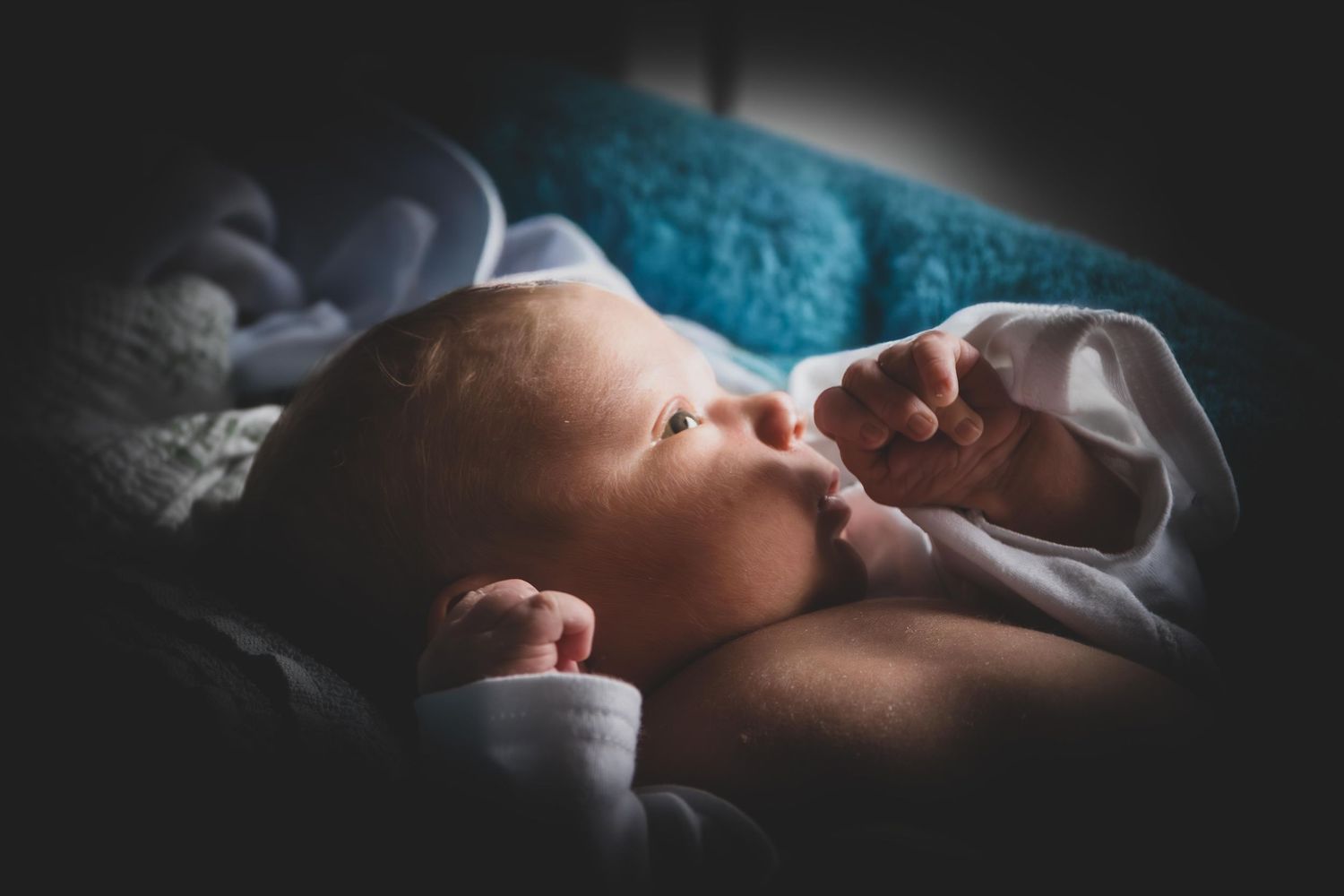 Newborn baby lying awake on a soft blanket in dim light, with tiny hands raised near face.