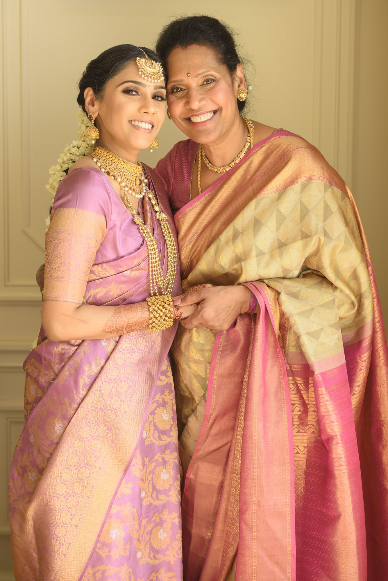 South Indian bride in lavender silk saree with gold jewelry poses with smiling mother in gold saree.