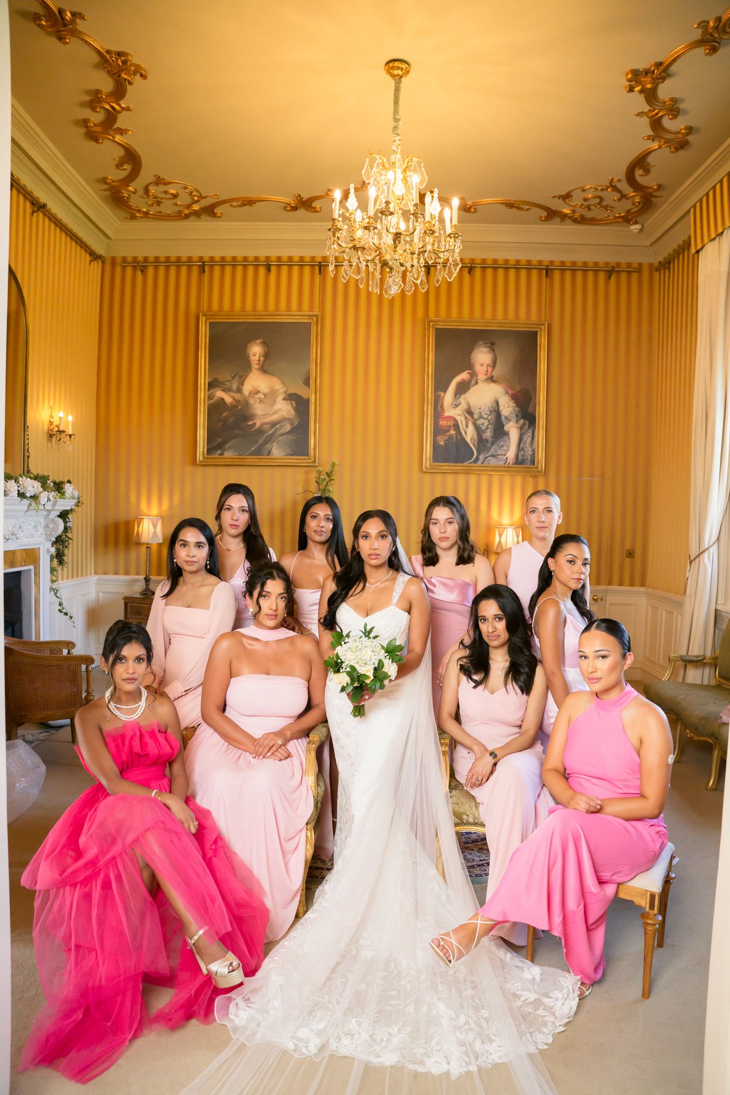 Bride in white gown poses with bridesmaids in pink dresses in an elegant gold-adorned room with chandelier.