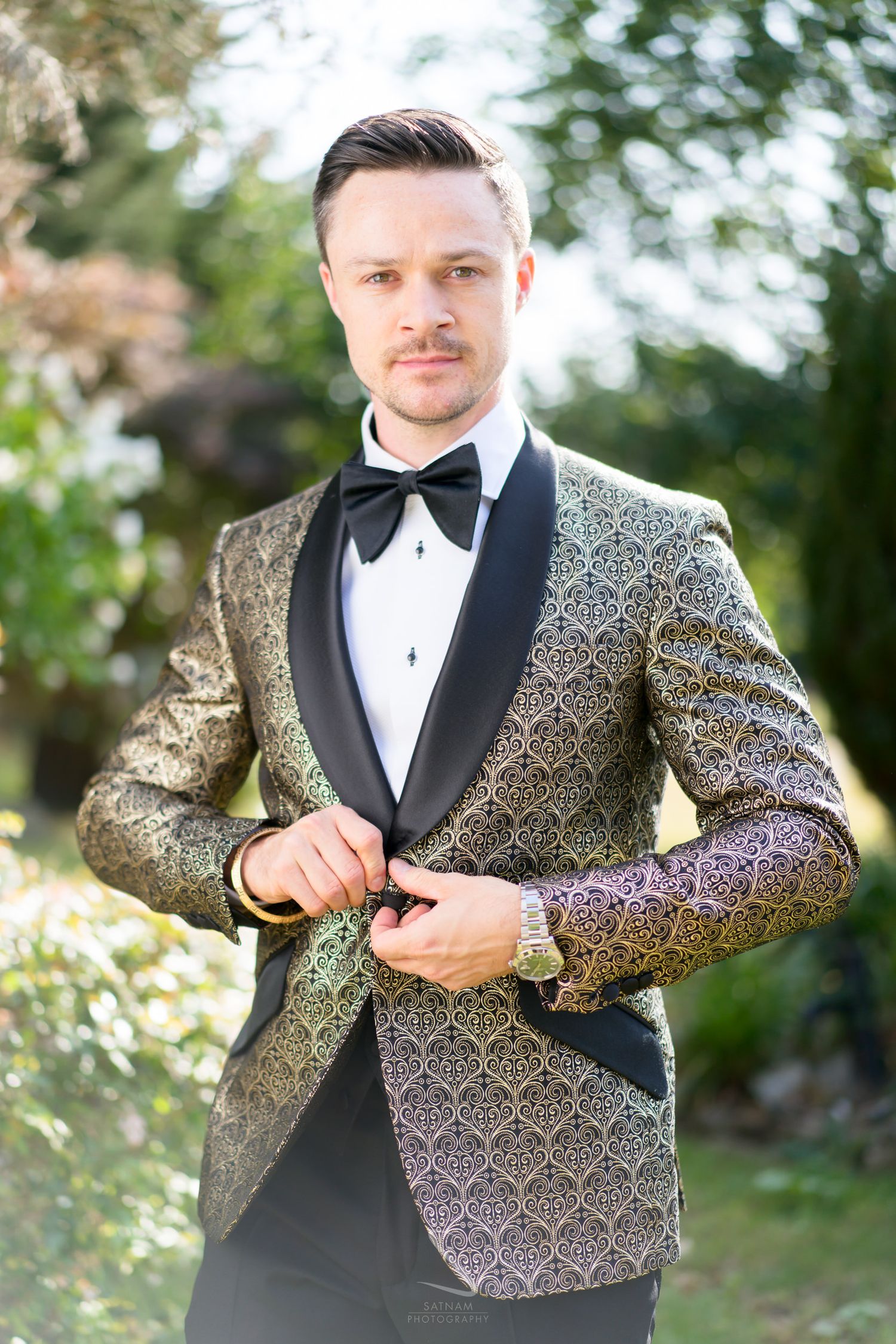 Groom in gold brocade tuxedo jacket with black bow tie posing outdoors at wedding.