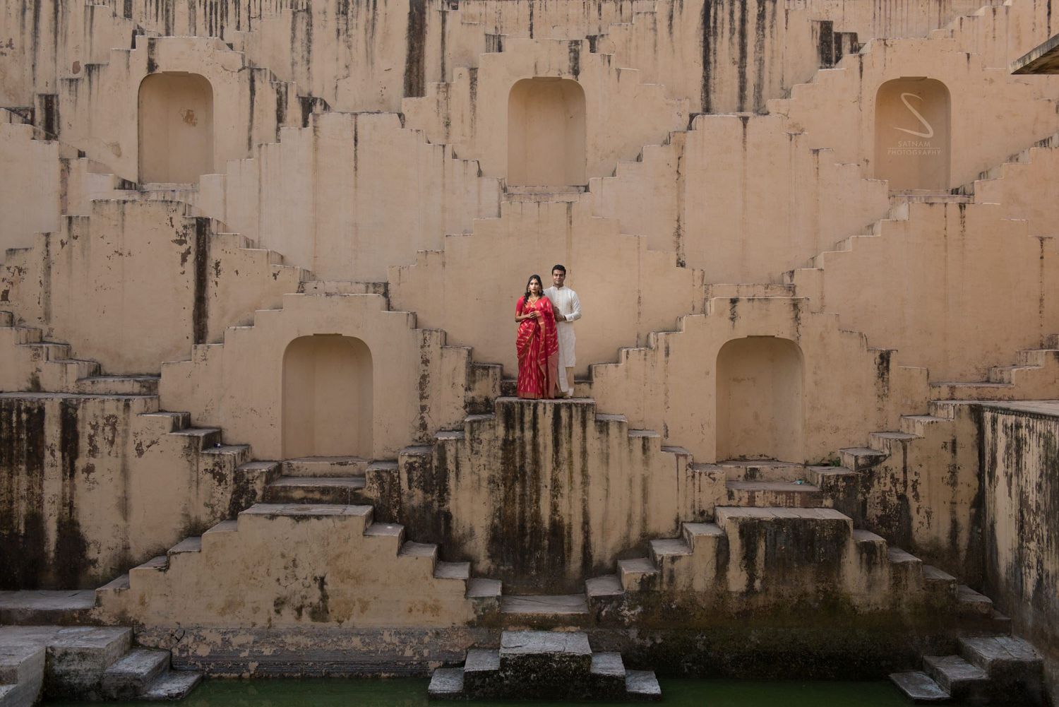 Couple posing on ancient stepwell architecture in Jaipur, woman in red saree and man in traditional Indian attire.