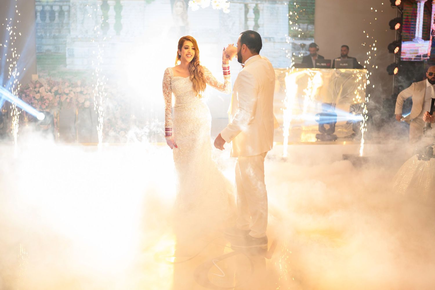Bride in white gown and groom share first dance surrounded by dramatic fog and stage lighting.
