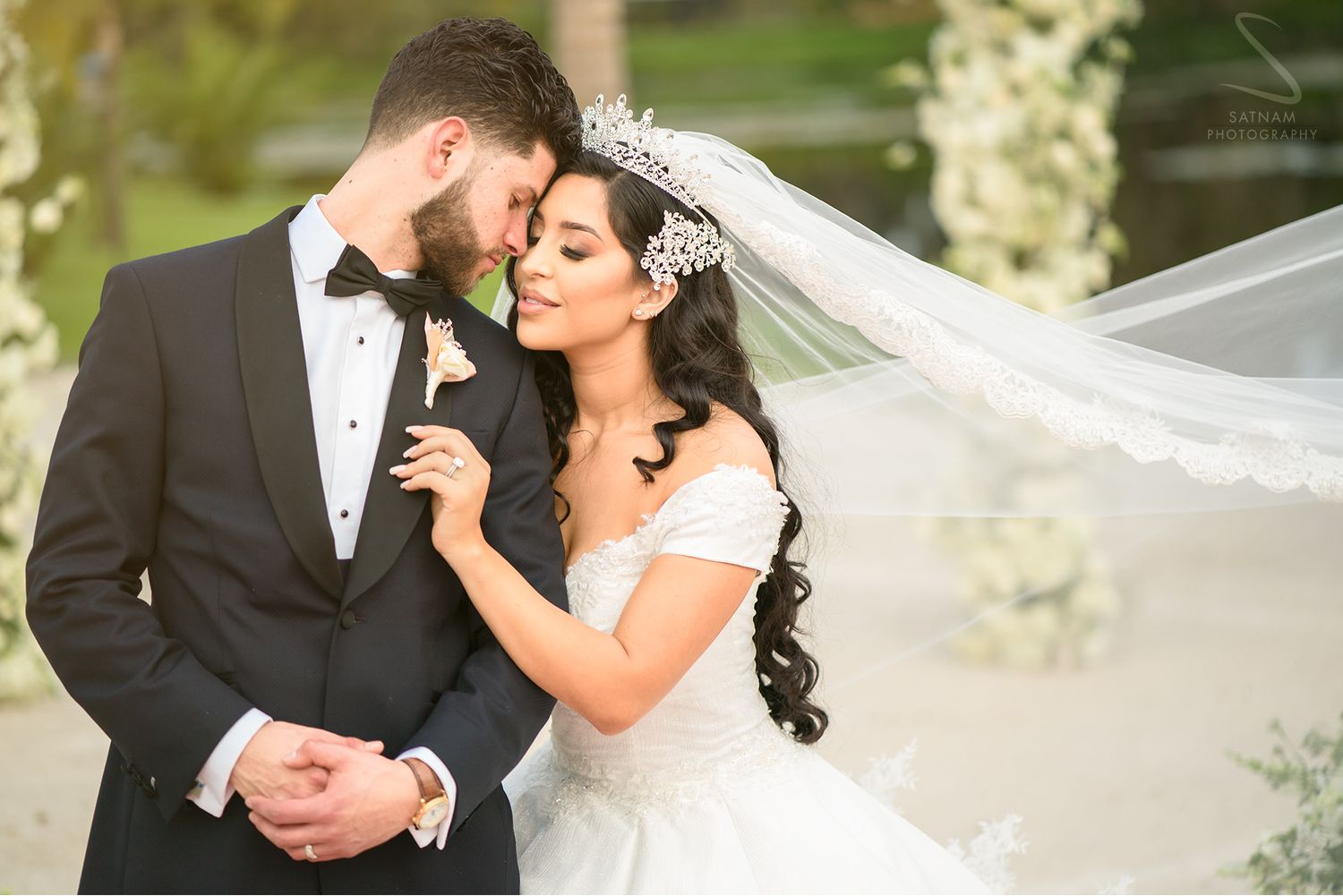 Bride in white gown with flowing veil embraces groom in black tuxedo at outdoor wedding ceremony.