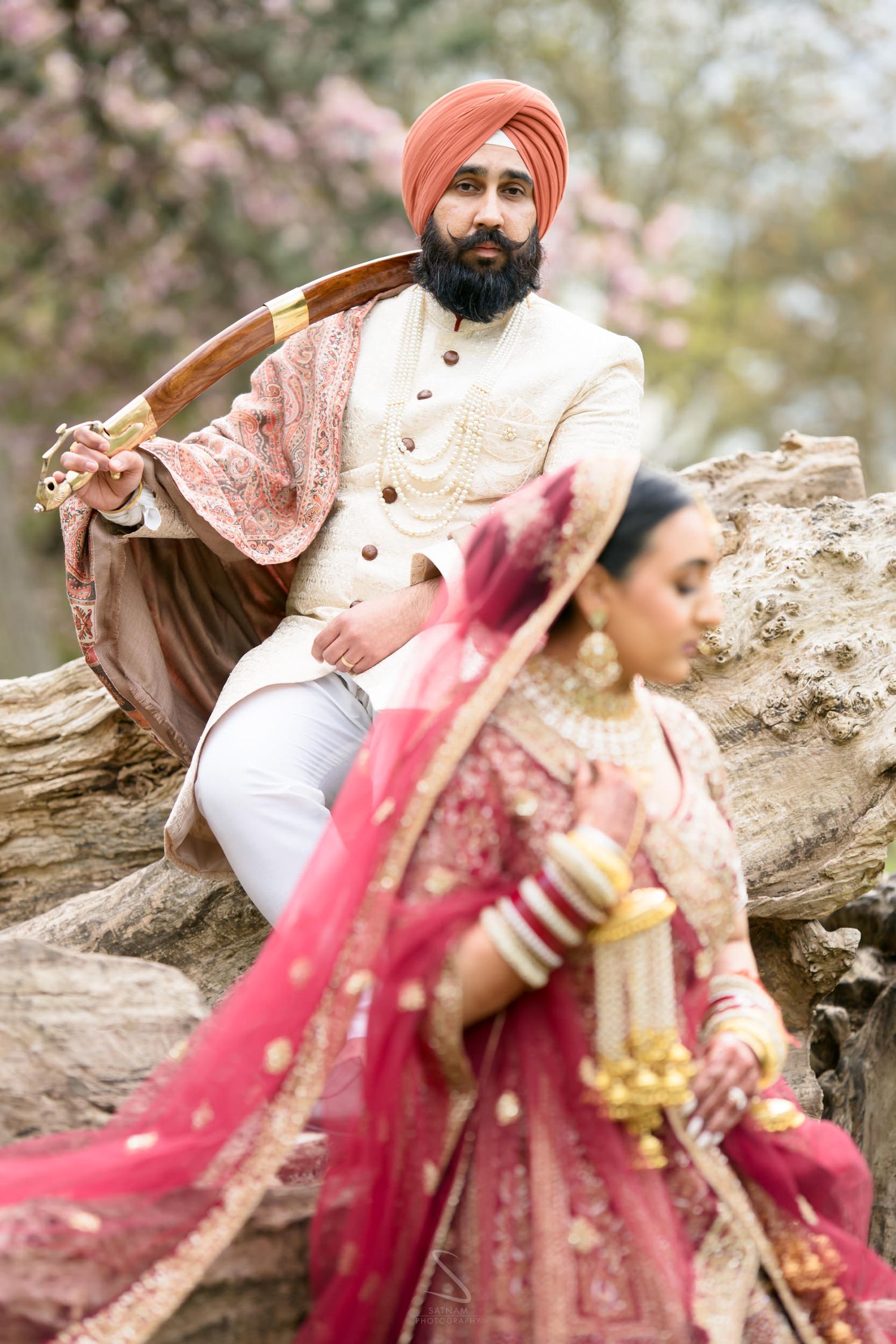 Sikh groom in orange turban sits on log holding kirpan while bride in red lehenga stands before him.