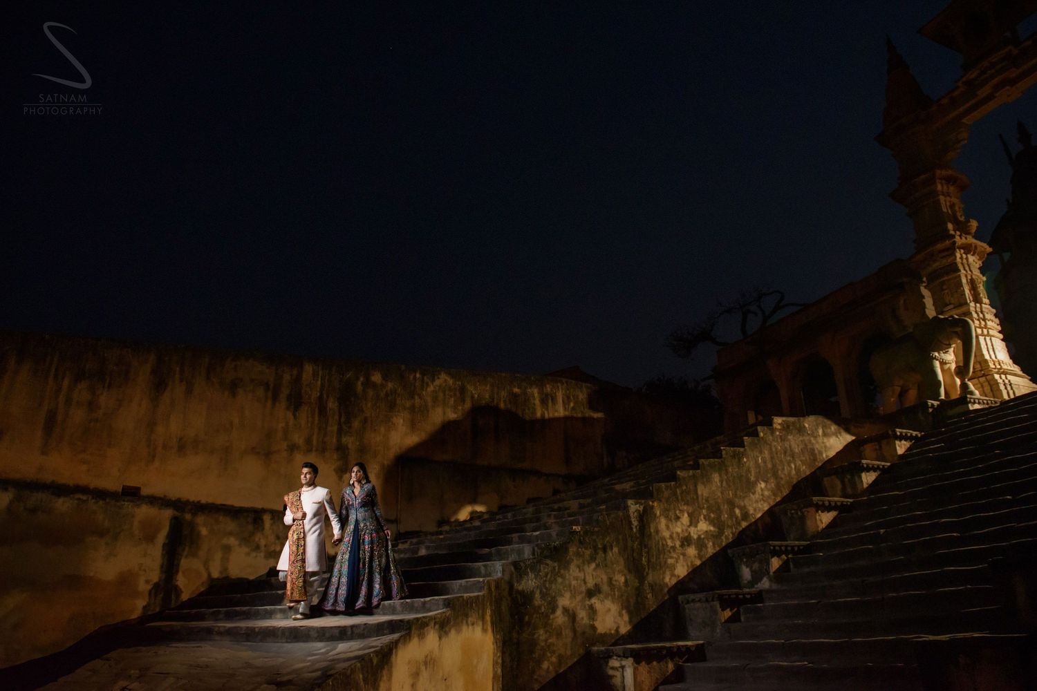 Couple descending ancient stone steps at illuminated historic monument in Jaipur at night.