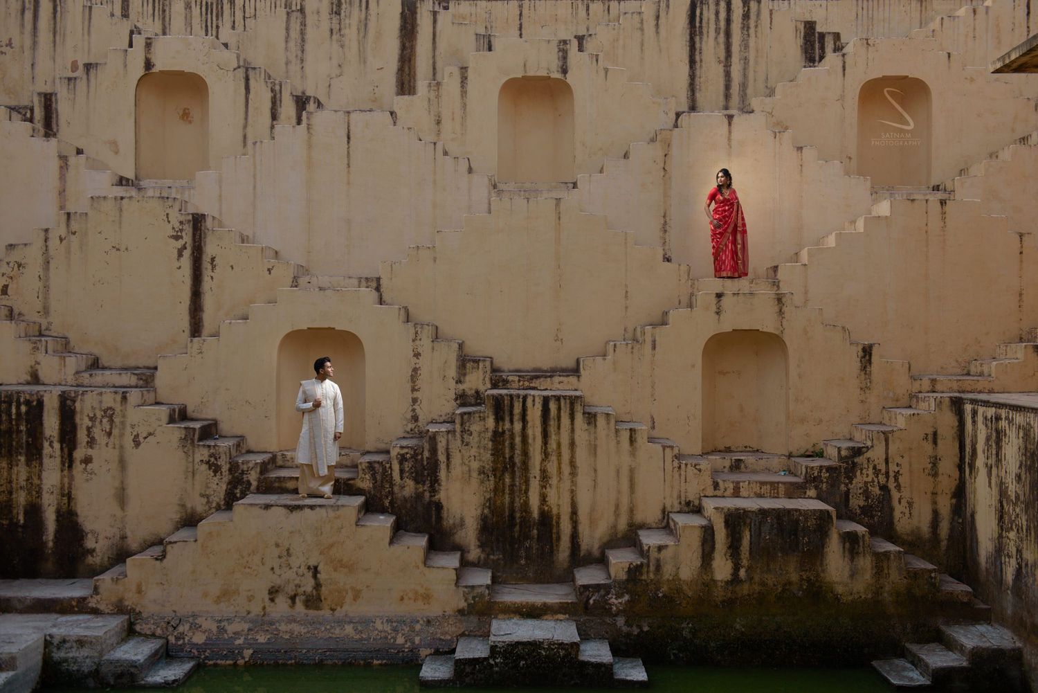 Indian couple in white and red traditional attire posing in geometric Panna Meena Ka Kund stepwell.