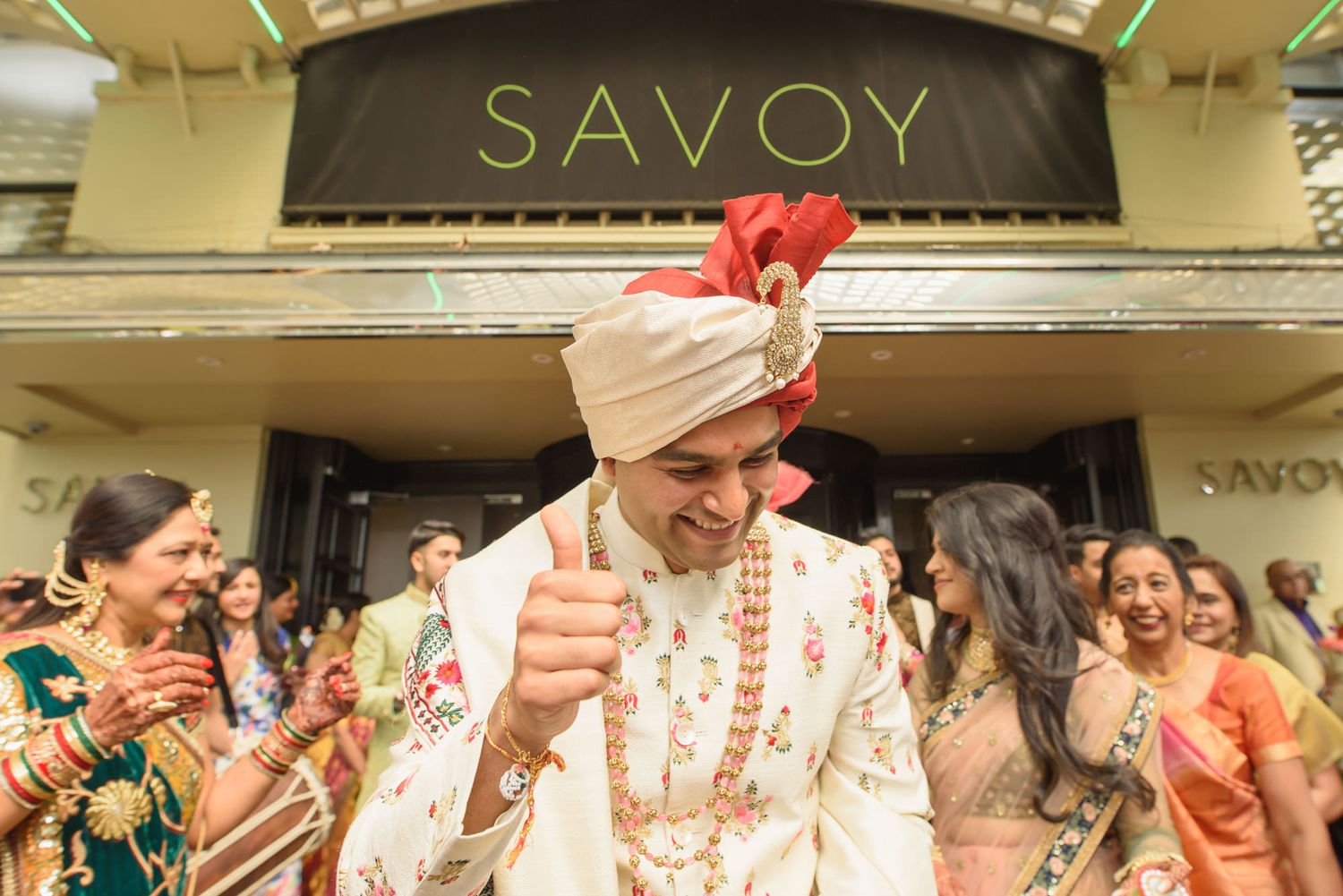 Cheerful Indian groom in white sherwani gives thumbs up outside the iconic Savoy hotel entrance.