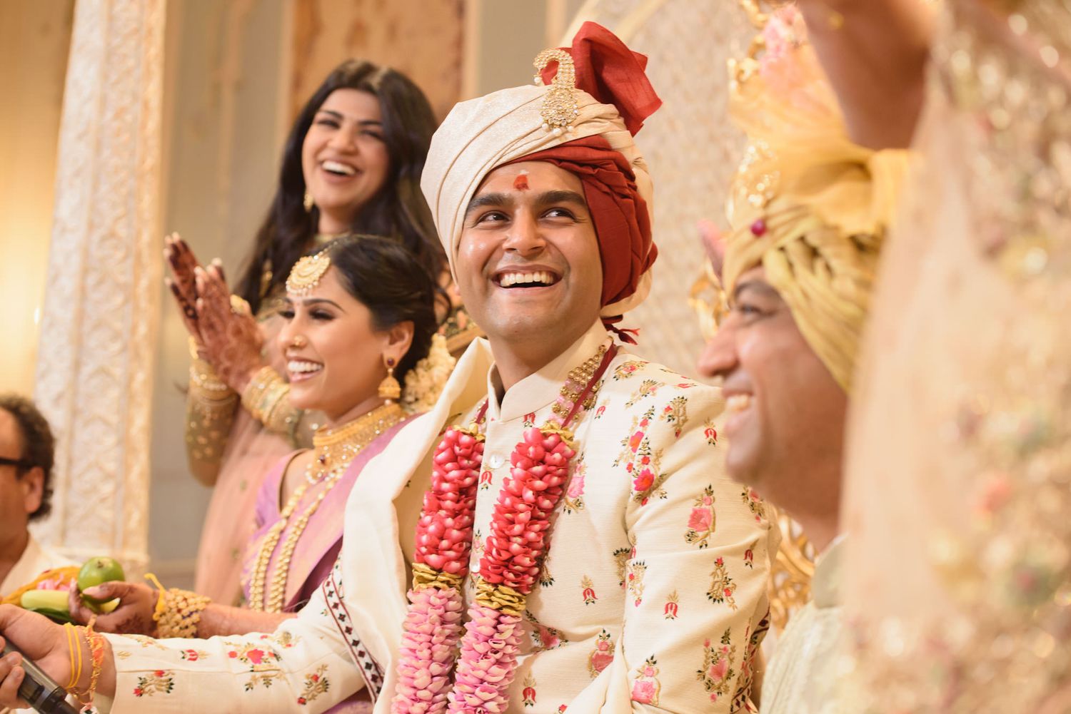 Smiling Indian groom in white floral sherwani and red turban celebrates joyfully at traditional wedding.