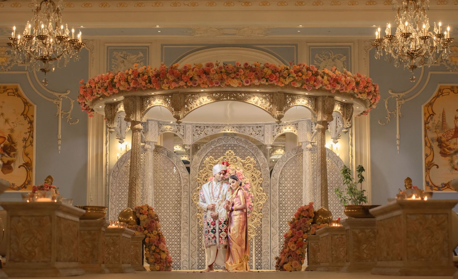 Indian bride and groom posing under ornate floral mandap with chandeliers at luxury wedding venue.