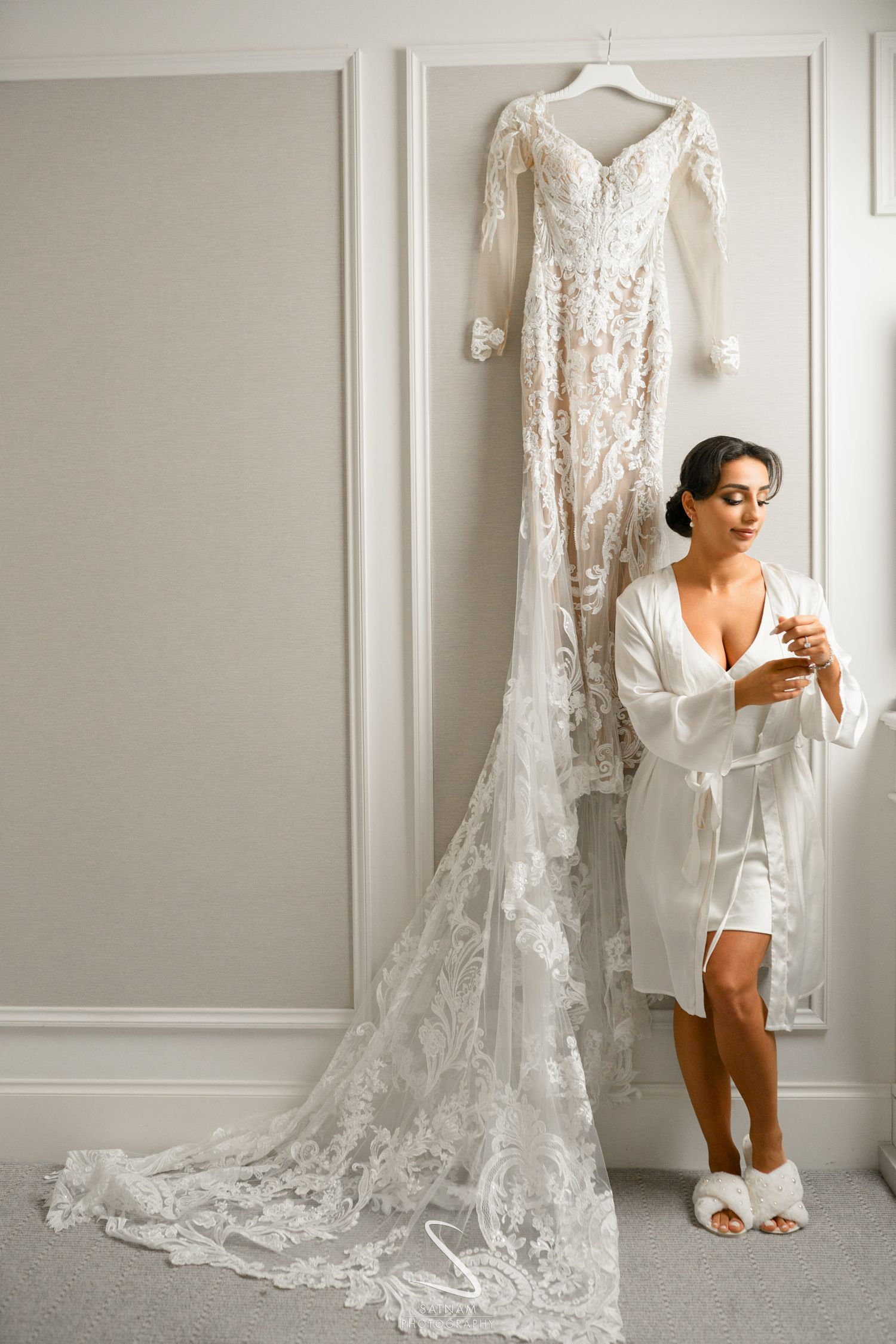 Bride in white robe admiring intricate lace wedding gown hanging on wall in bright dressing room.