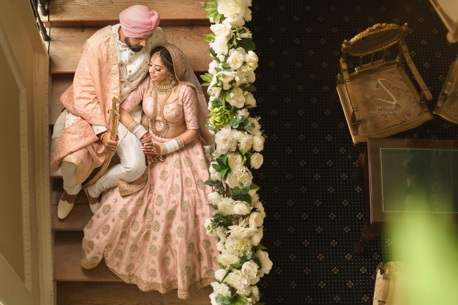 Sikh couple in matching blush pink outfits seated on floral decorated staircase at wedding.