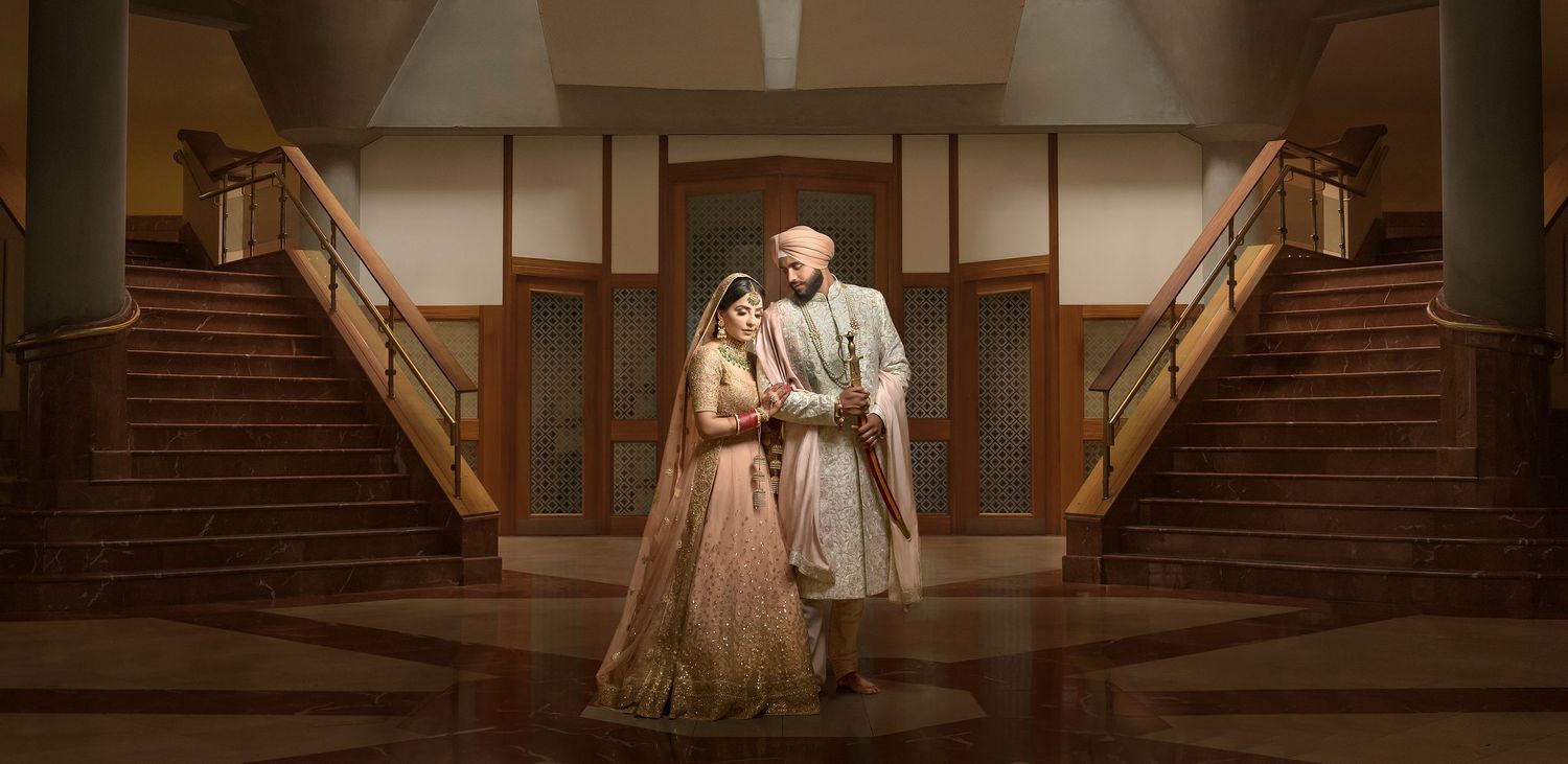 Indian bride and groom in traditional attire stand between grand staircases.