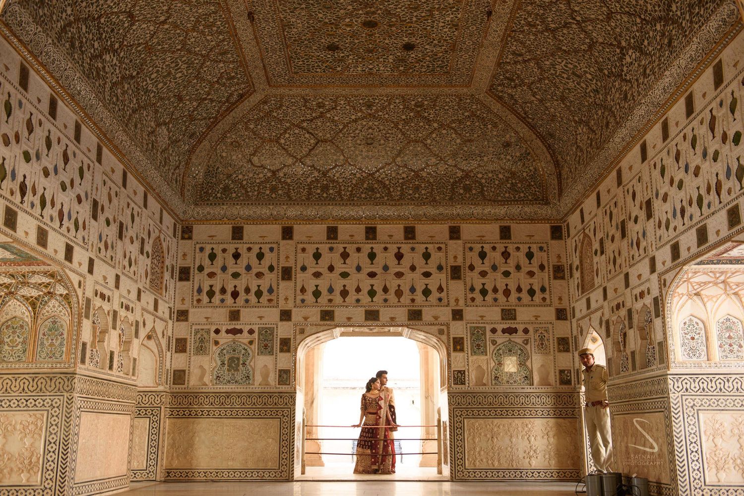 Bride in red lehenga stands beneath an elaborately decorated ceiling in Amer Fort, Jaipur.
