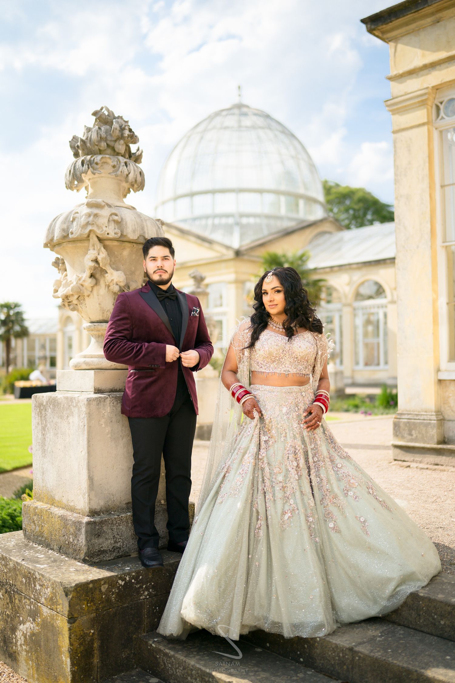Indian couple posing at grand greenhouse venue, bride in blush lehenga, groom in burgundy blazer.