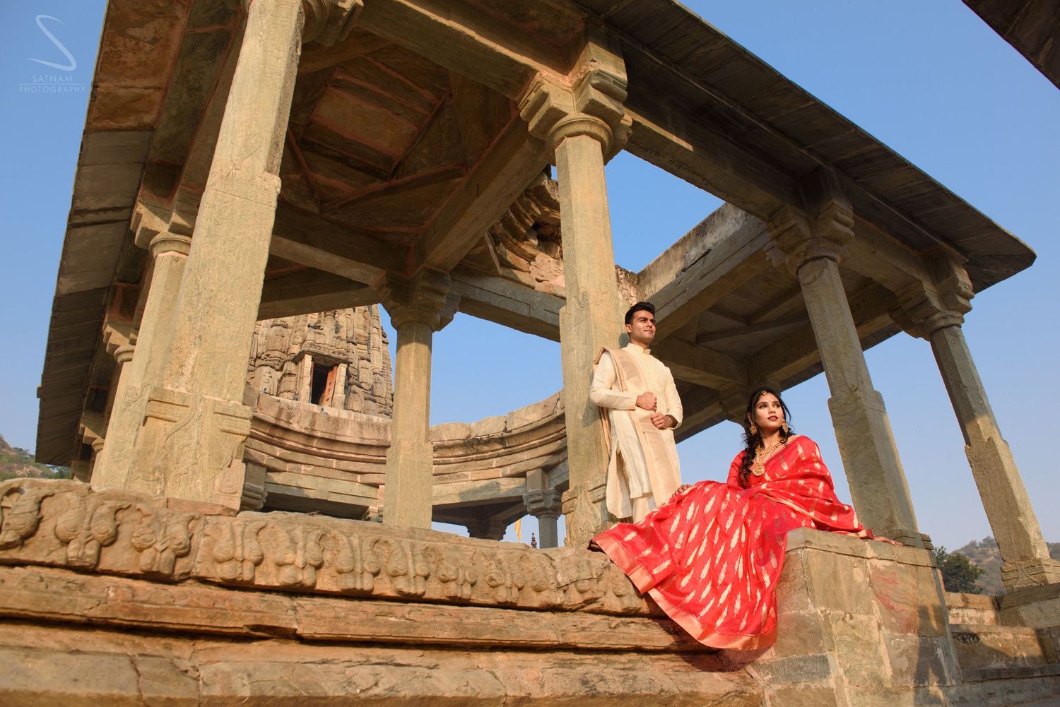 Couple in traditional Indian outfits posing at ancient stone temple ruins under blue sky.