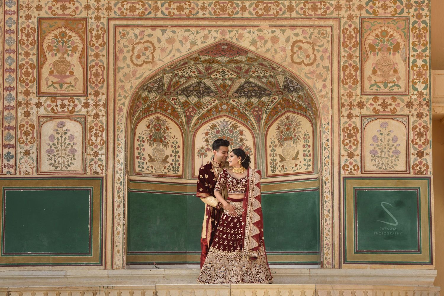 Indian couple in maroon wedding outfits embraces beneath an ornate painted arch at Amer Fort.