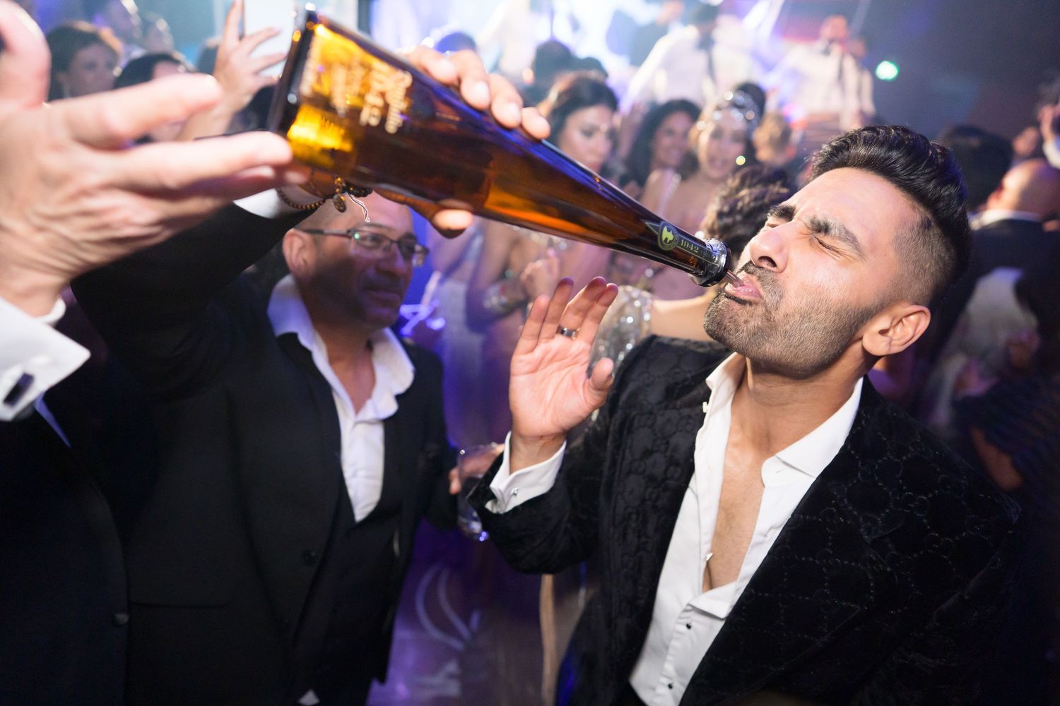 Man drinks champagne from bottle at lively wedding reception party with crowd in background.