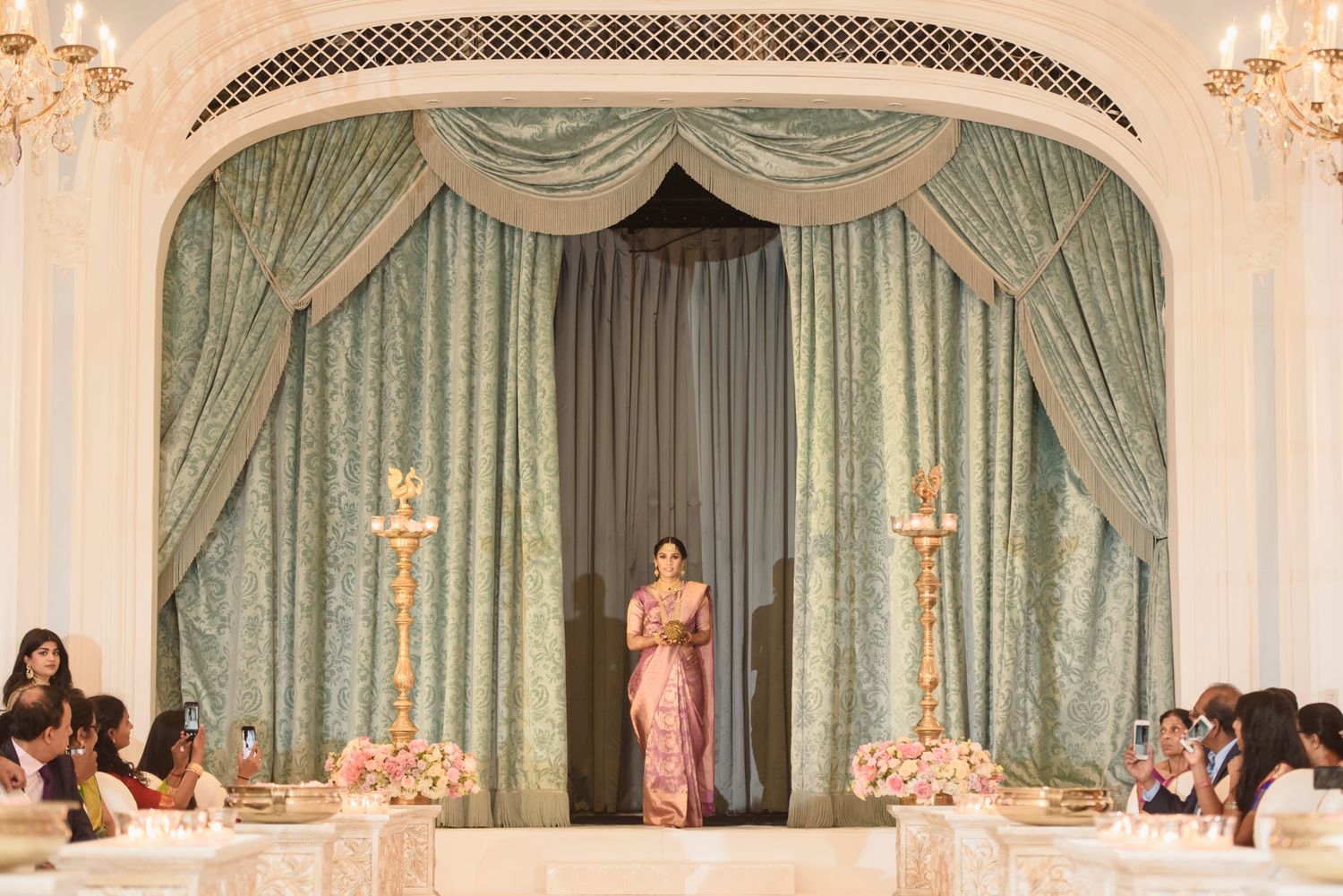 Elegant woman in pink saree stands at the entrance of a grand Indian wedding venue with teal drapes.
