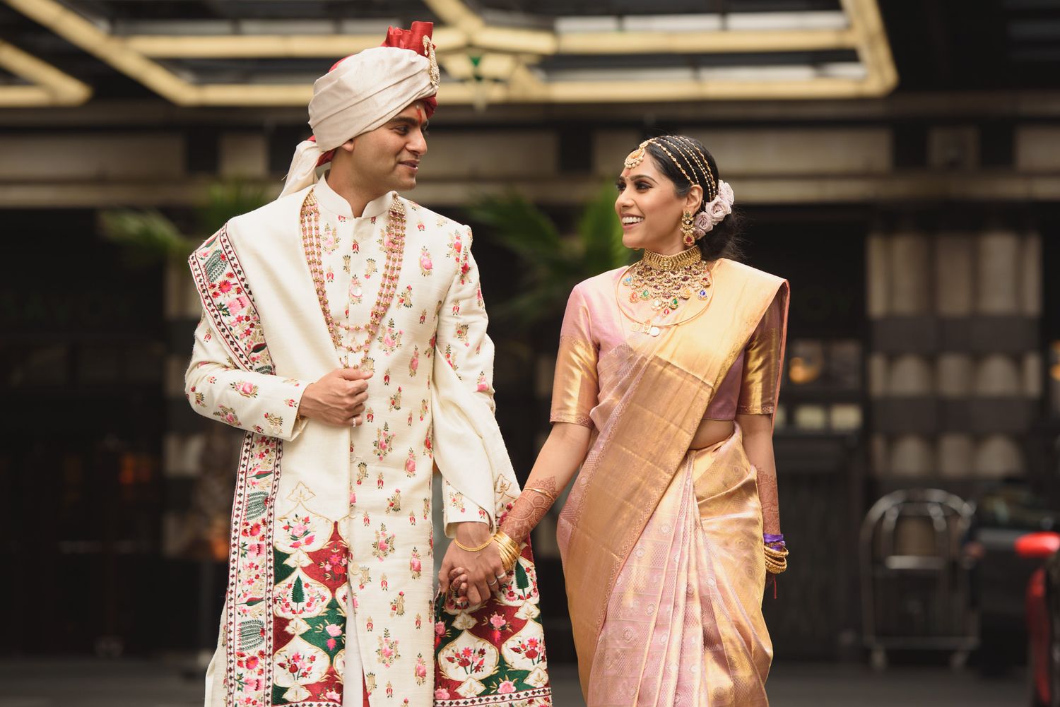 Indian bride in pink silk saree and groom in floral sherwani holding hands at wedding.
