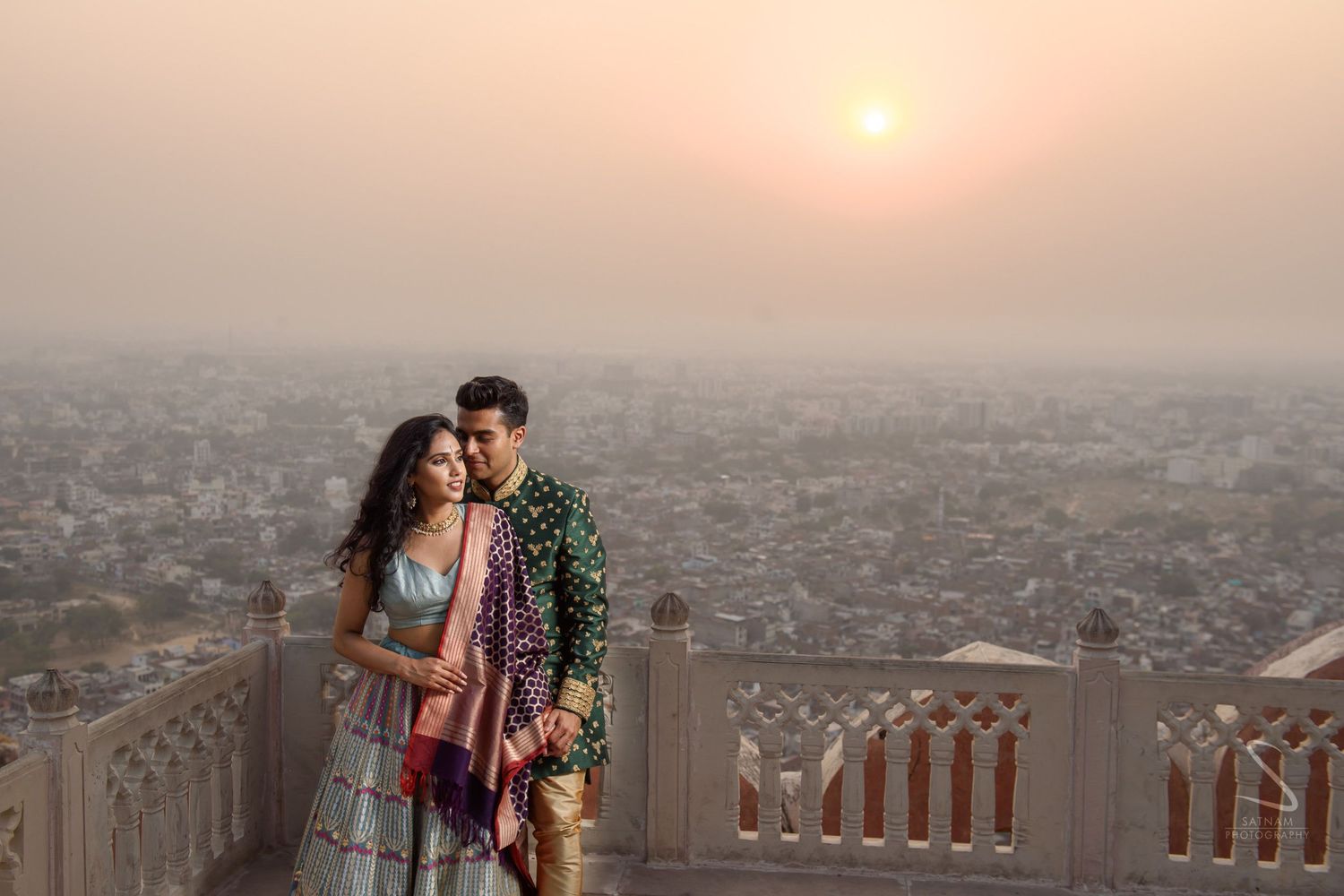 Indian couple embracing at sunset overlooking Jaipur cityscape, dressed in traditional wedding attire.
