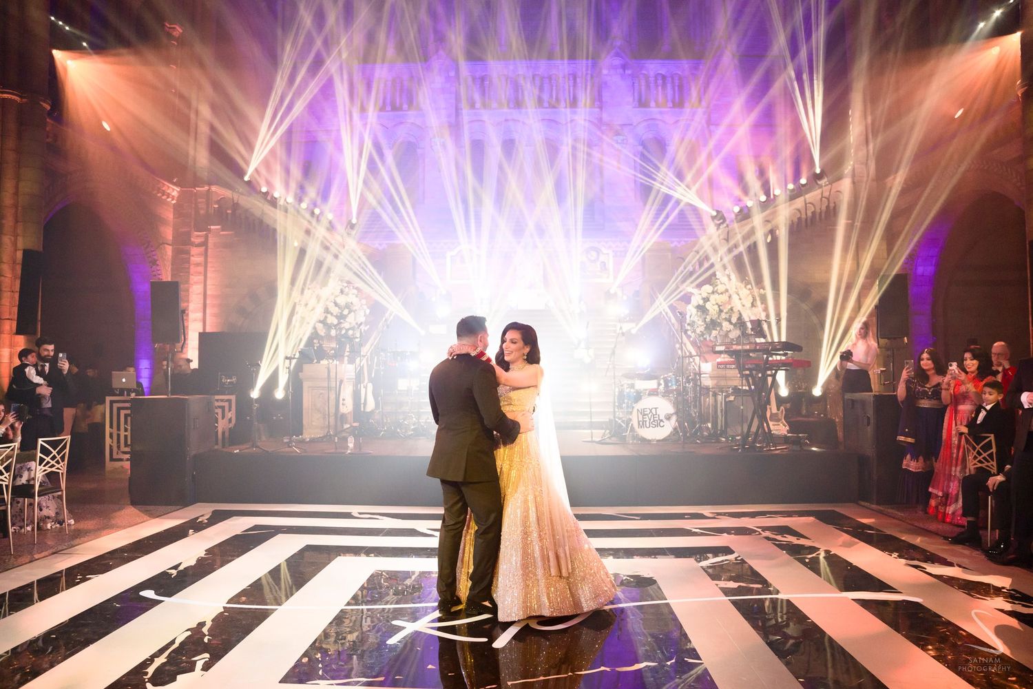 Couple sharing first dance on checkered floor with dramatic stage lighting and live band at wedding reception.