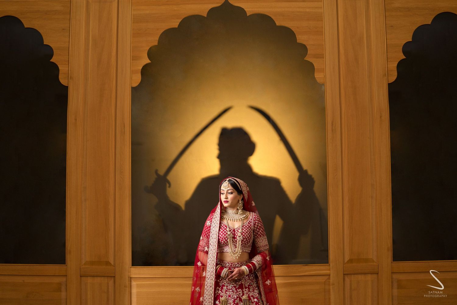 Indian bride in red bridal lehenga posing with dramatic warrior shadow projection on ornate wooden backdrop.