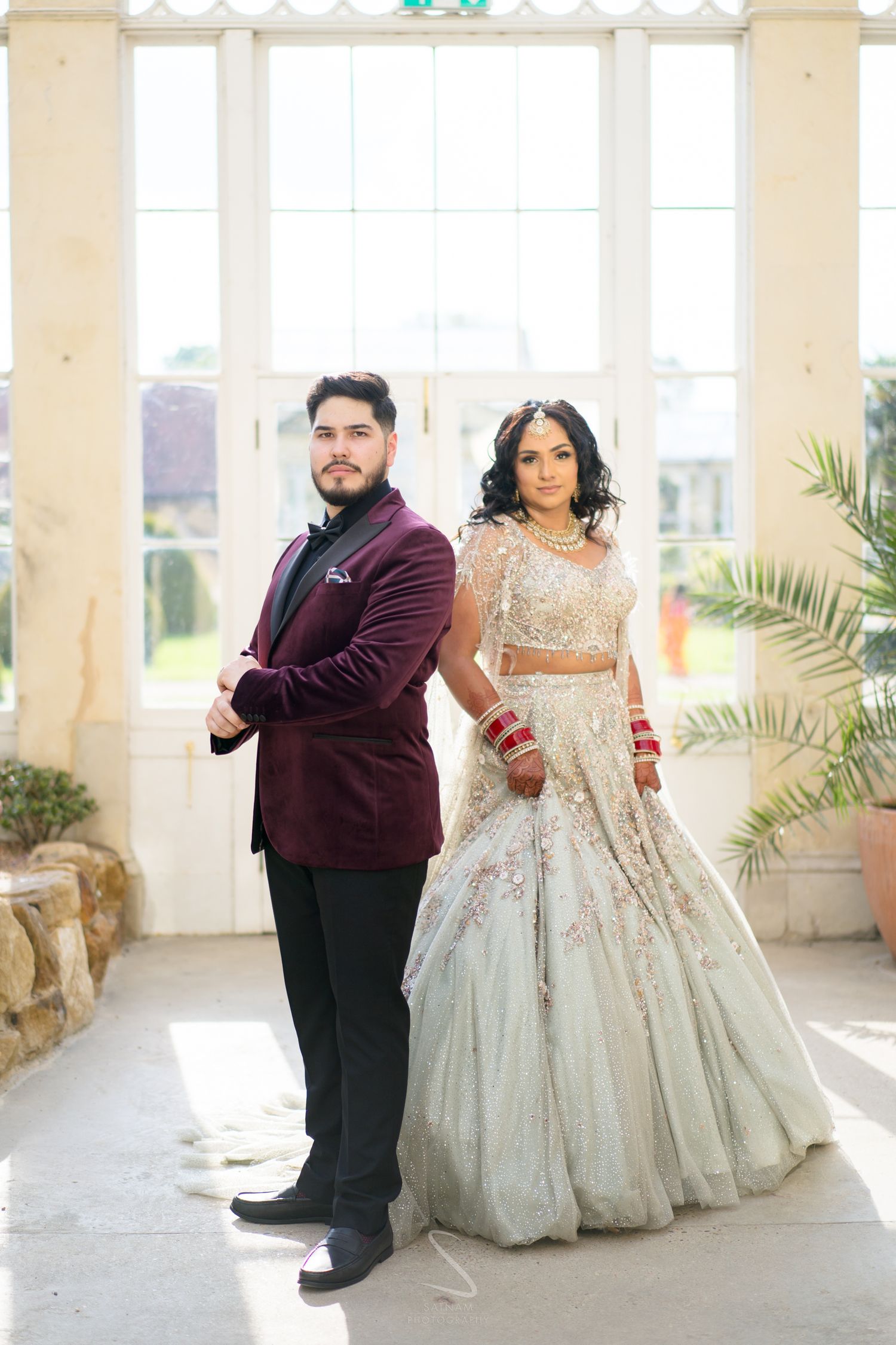 An Indian couple poses back to back, the bride in a silver lehenga and groom in a burgundy velvet blazer.