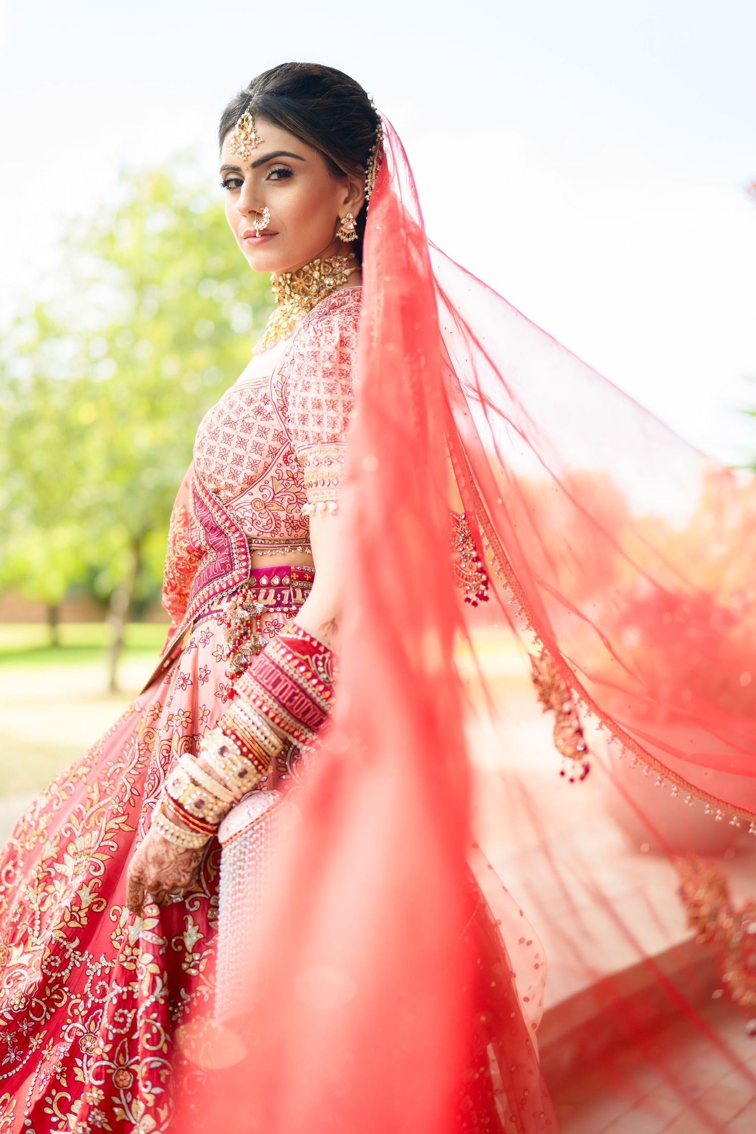 Indian bride in red embroidered lehenga poses with flowing red veil billowing in the wind outdoors.