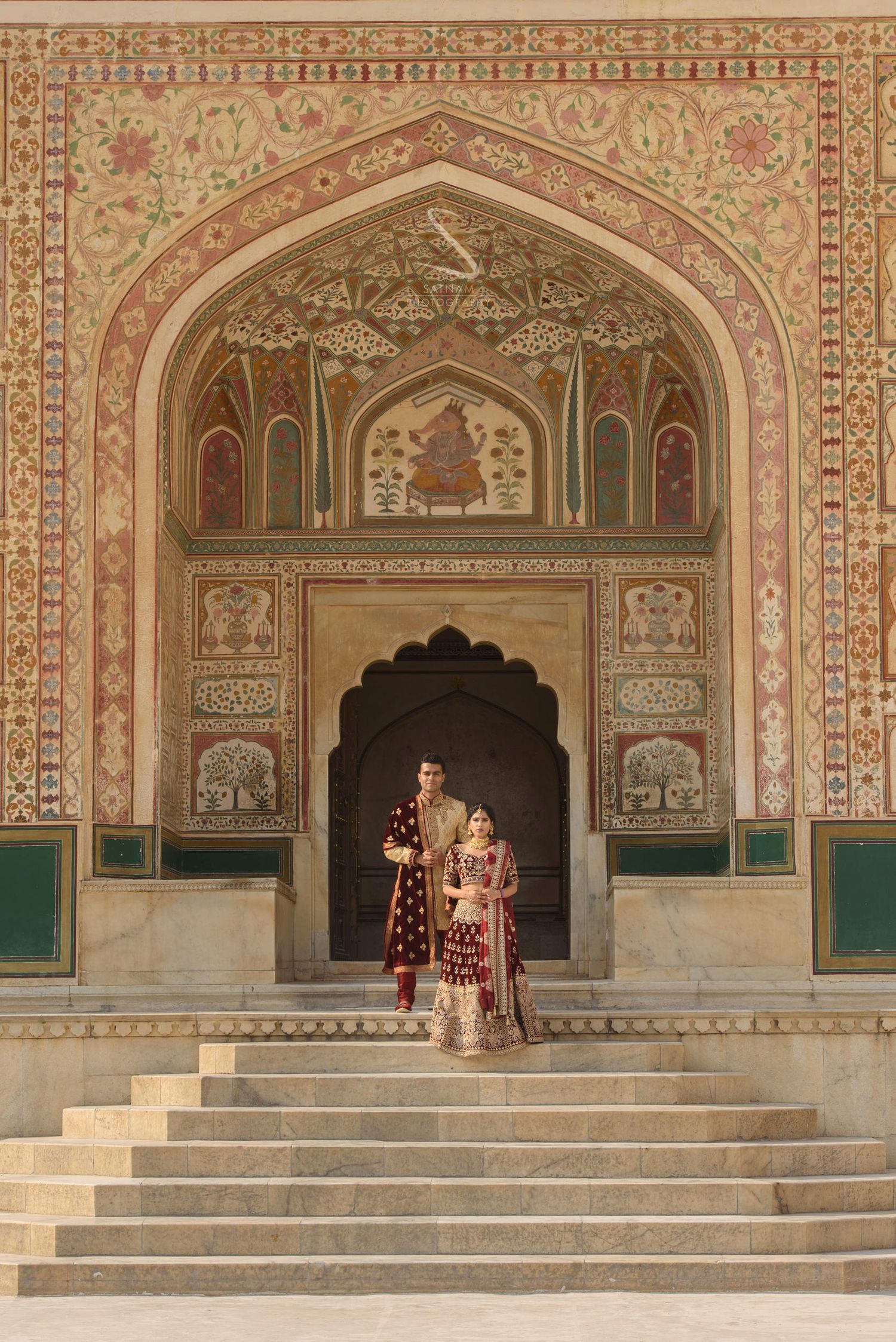 Indian wedding couple in maroon bridal attire standing under elaborately painted Mughal archway at Amer Fort.