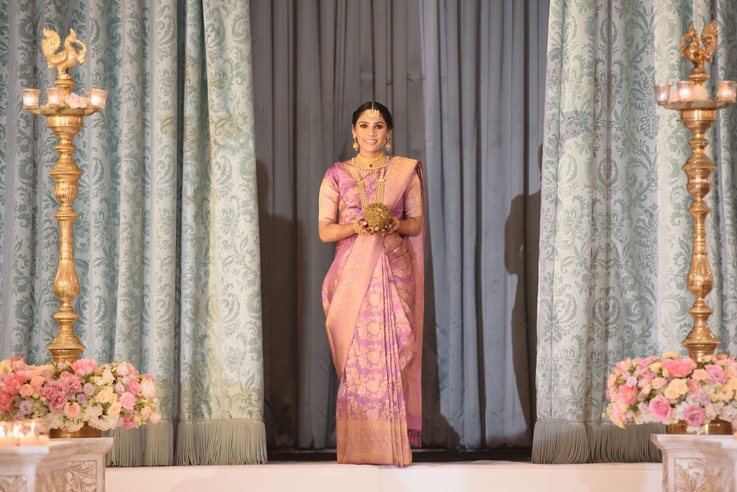 Indian woman in pink silk saree with gold jewelry stands gracefully at a decorated wedding venue.