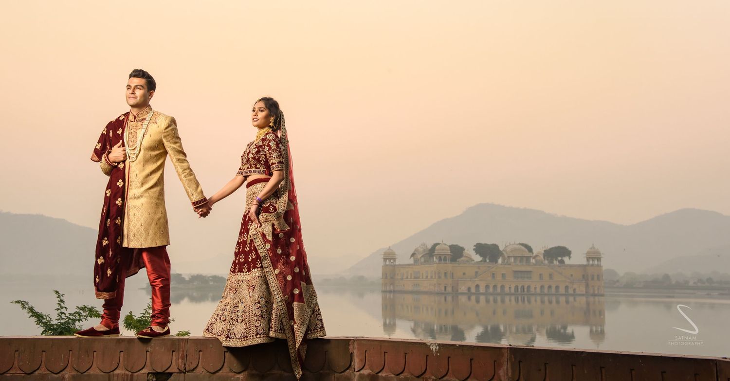 Indian bride and groom in maroon bridal outfits holding hands at Jal Mahal lake palace, Jaipur at dusk.
