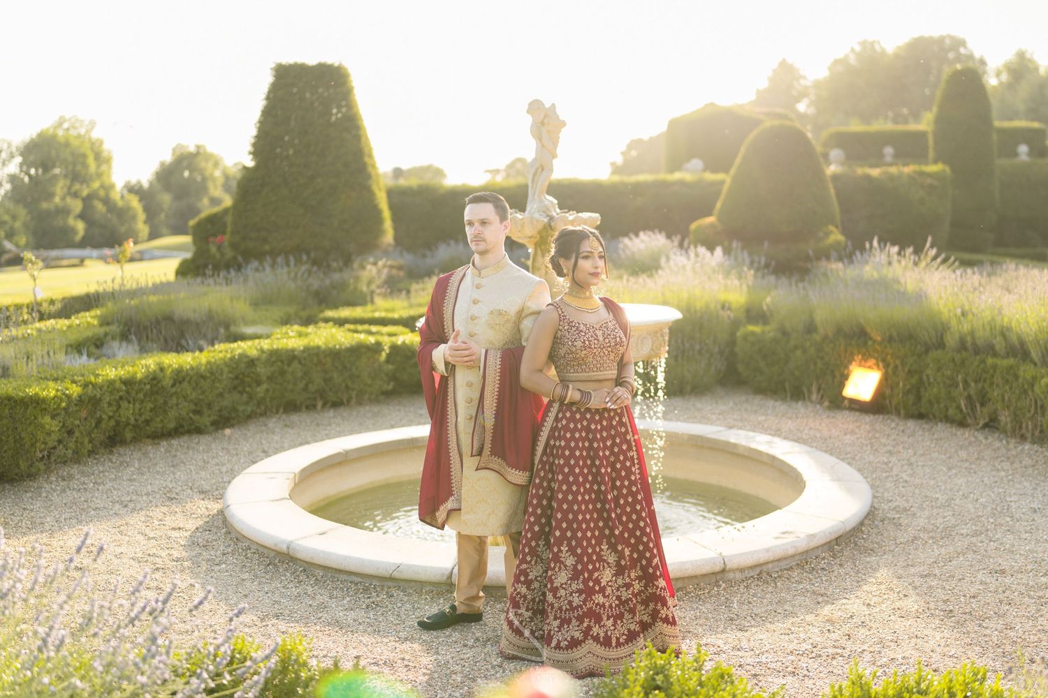 Intercultural wedding couple in traditional Indian attire poses by stone fountain in formal garden.