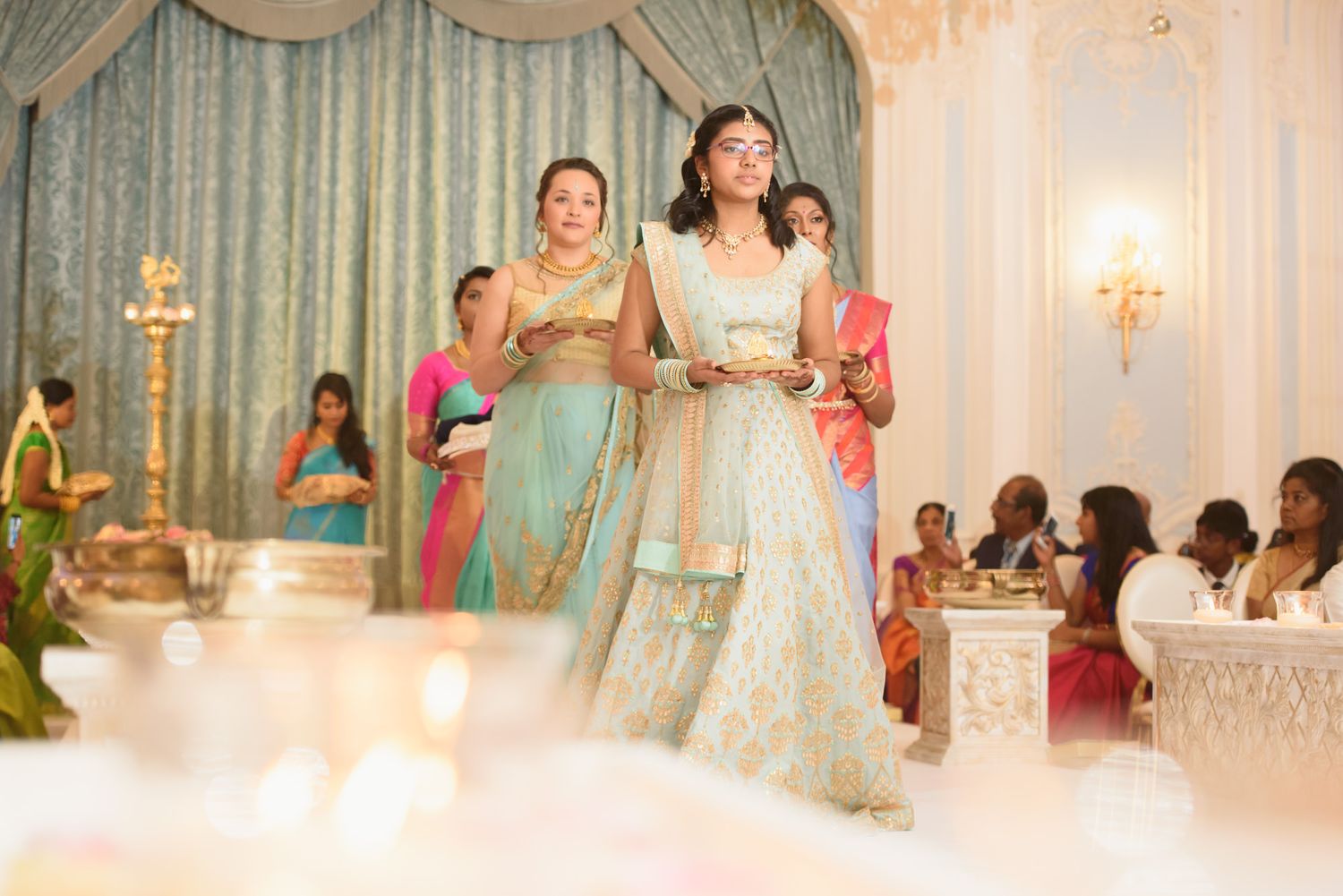 Women in colorful Indian attire carry ceremonial items during a traditional Hindu wedding ritual.