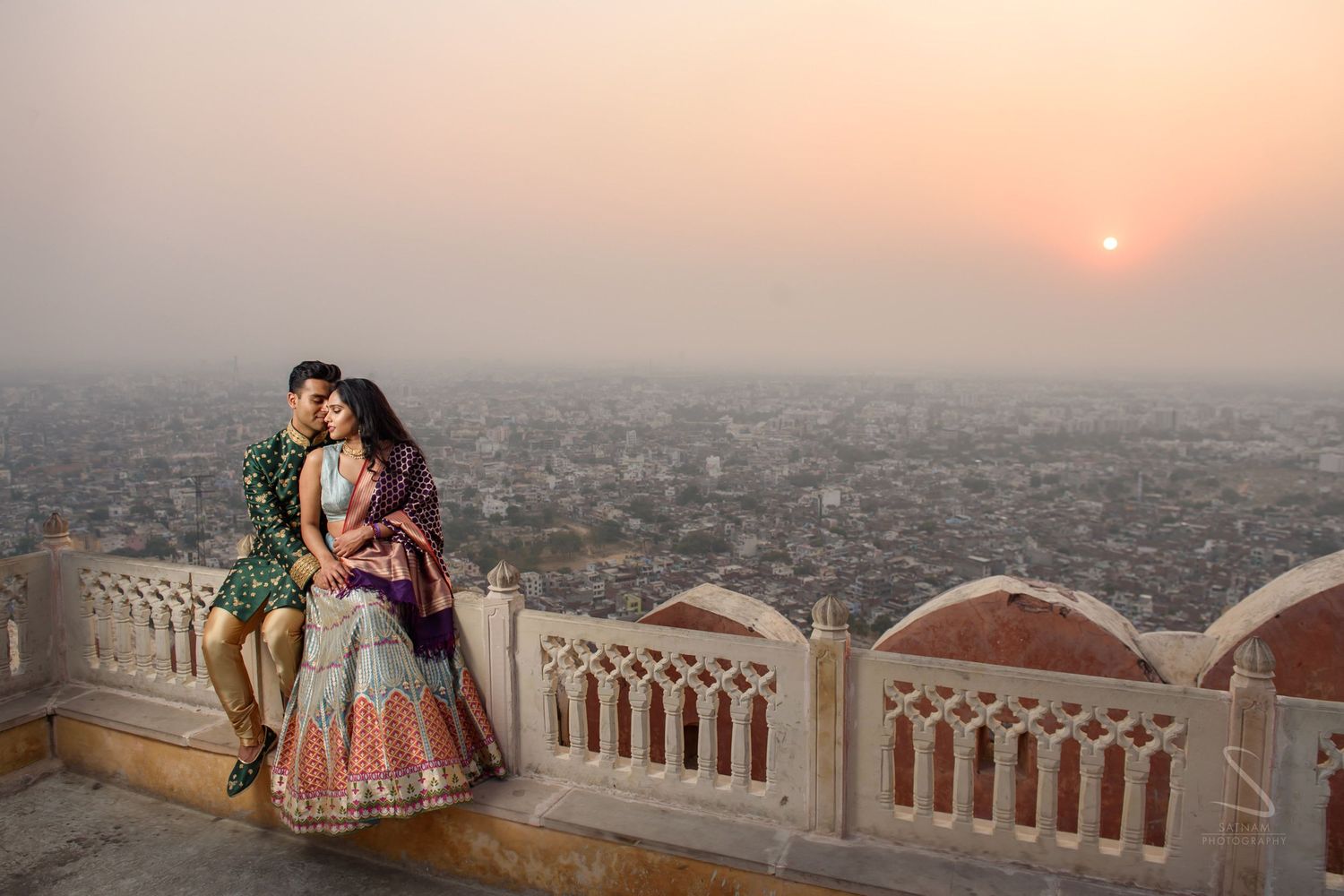 Indian couple in bridal lehenga and sherwani embracing at sunset overlooking Jaipur cityscape.
