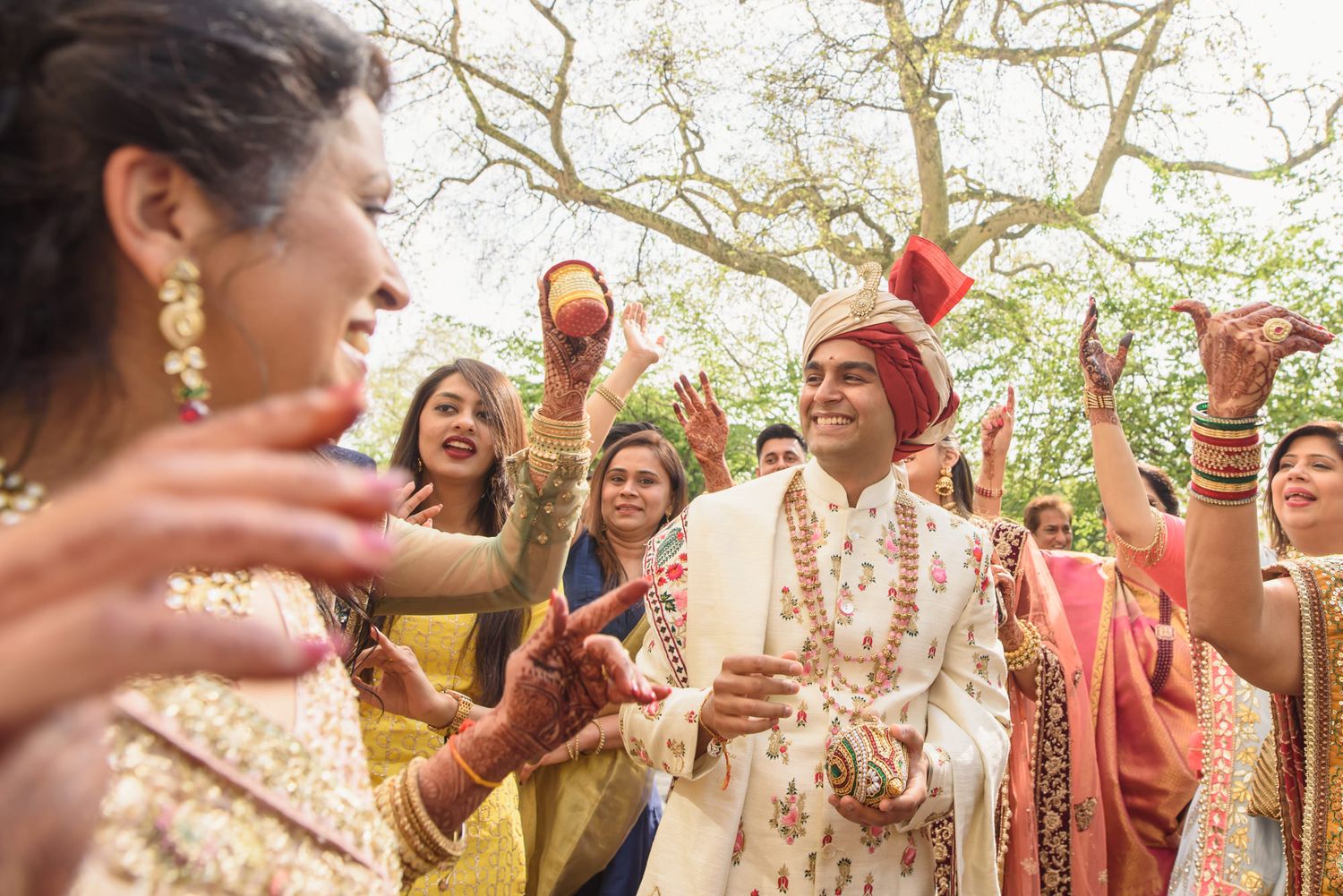 Smiling Indian groom in white sherwani celebrates with joyful wedding guests during outdoor baraat.