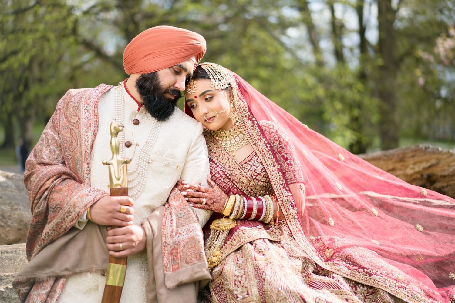 Sikh couple in pink and cream traditional bridal attire share tender moment seated on rustic log.