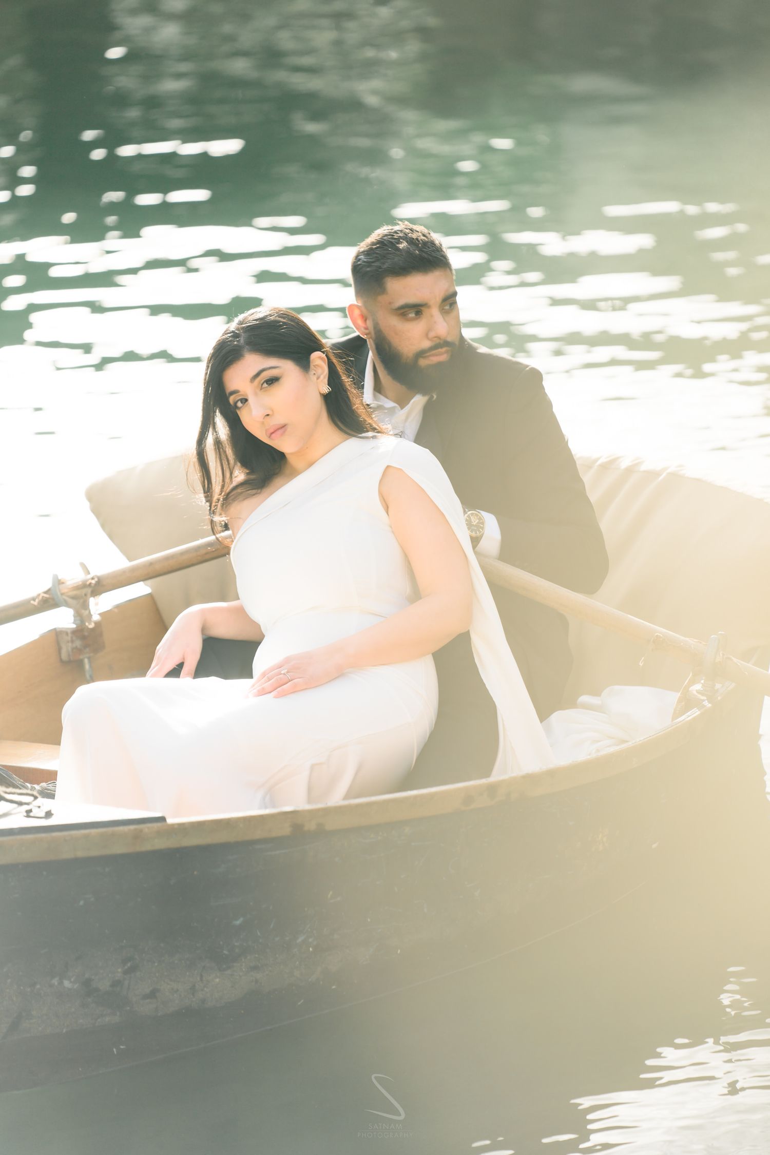 A couple in wedding attire sits in a wooden rowboat on calm water, the bride in white and groom in a dark suit.