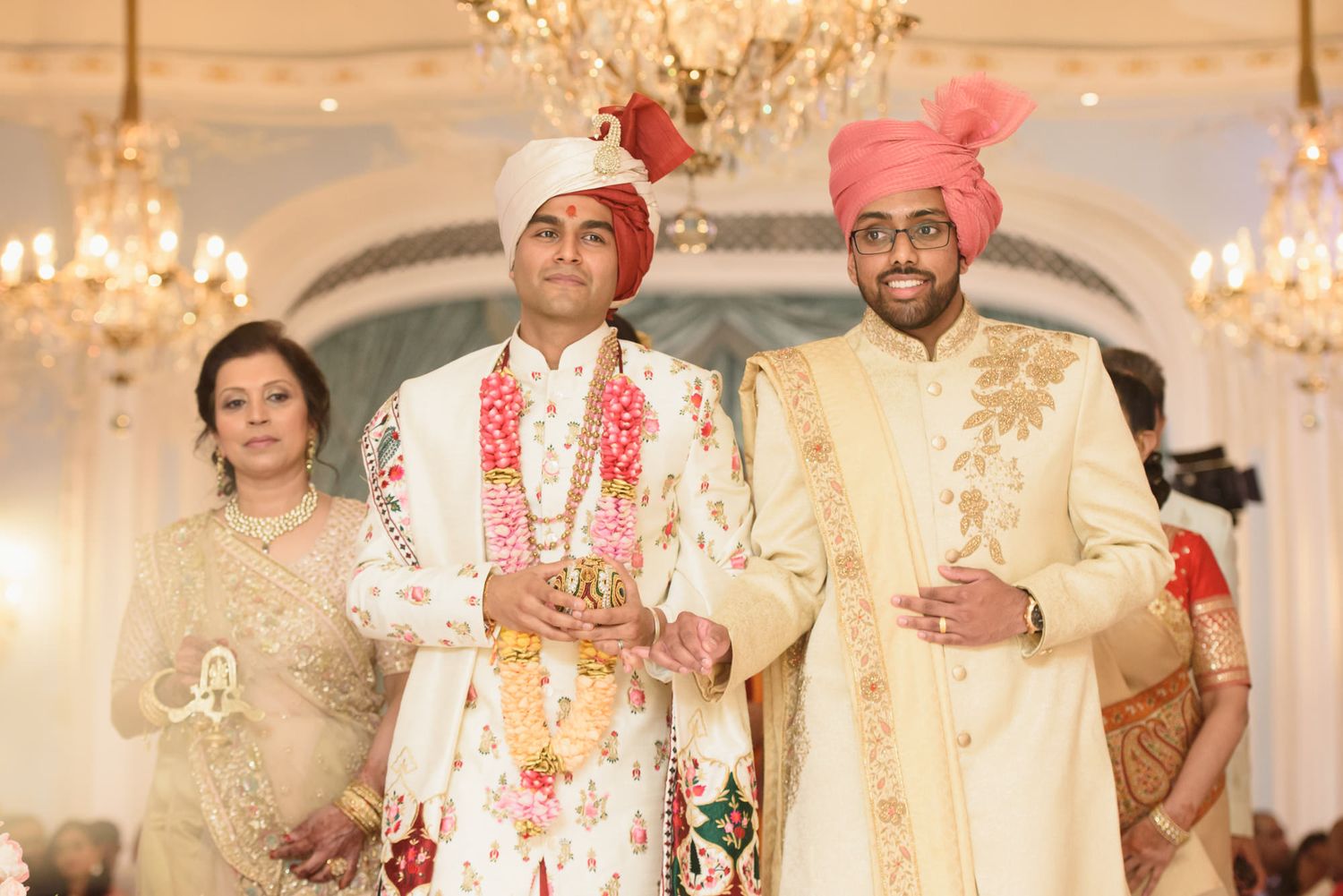 Three people in traditional Indian wedding attire walk down the aisle at an elegant Hindu wedding ceremony.