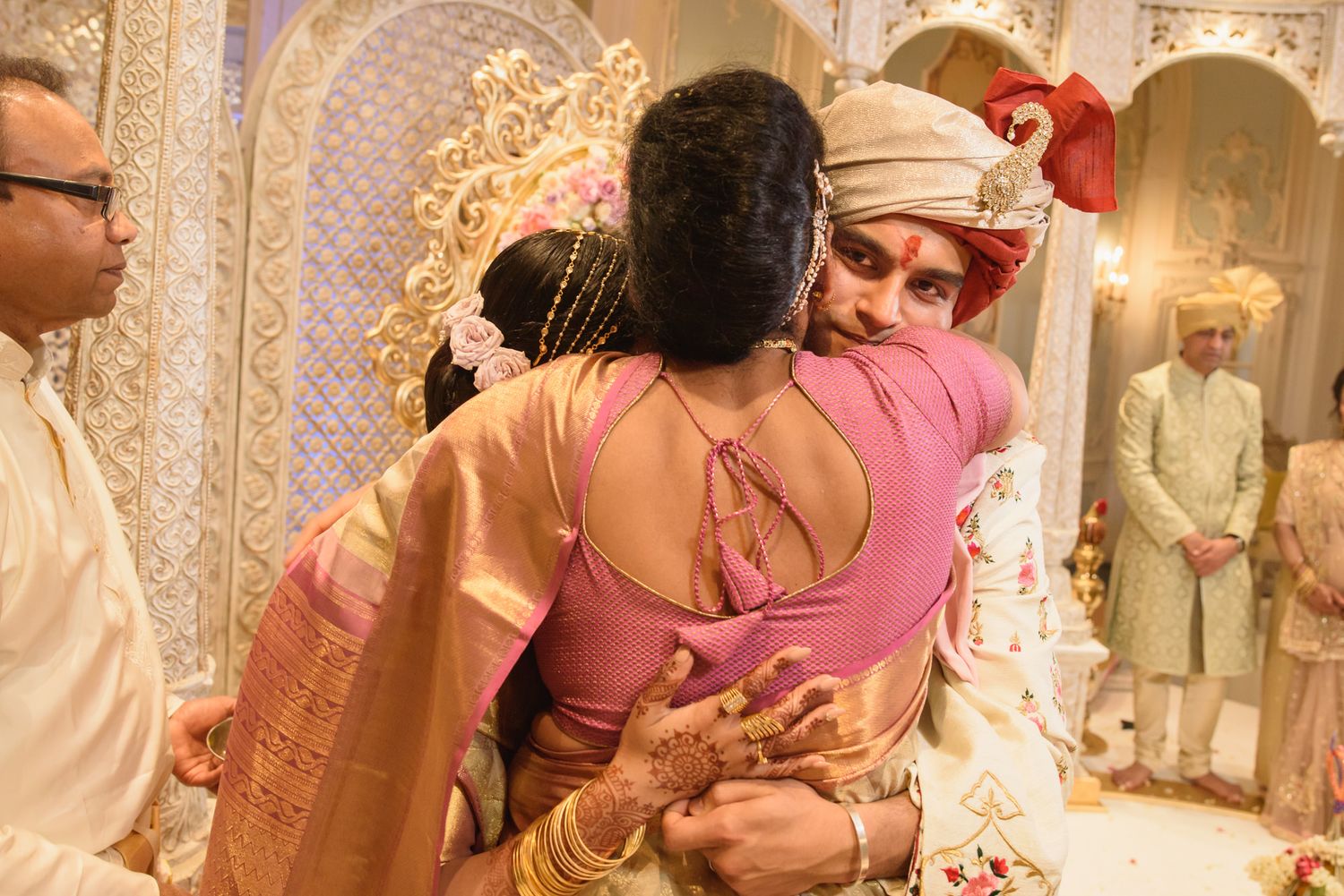 Groom in embroidered sherwani sharing warm embrace with woman in pink saree at Indian wedding.